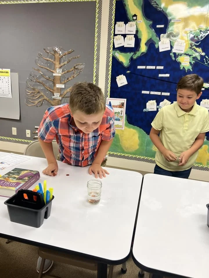 Two boys participating in a classroom activity involving a glass of water and objects on a table. One boy is leaning over the table, looking into the glass. The classroom features a large map on the wall.