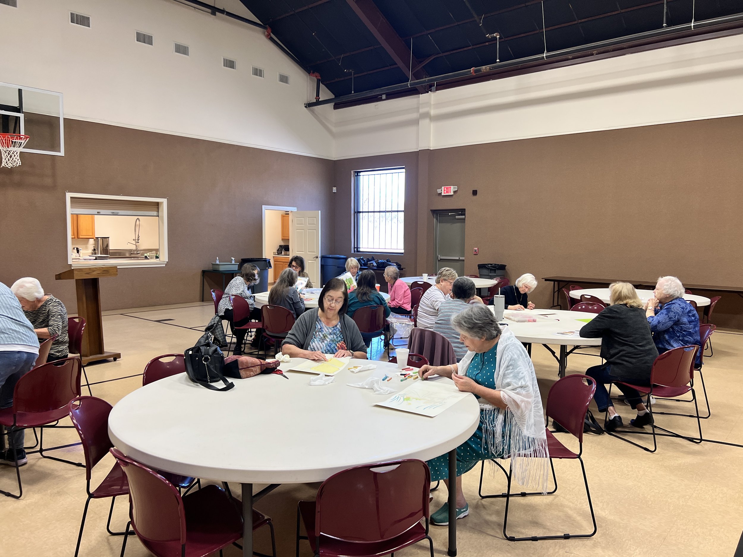 Group of elderly women sitting at round tables in a community hall, engaging in arts and crafts activities.