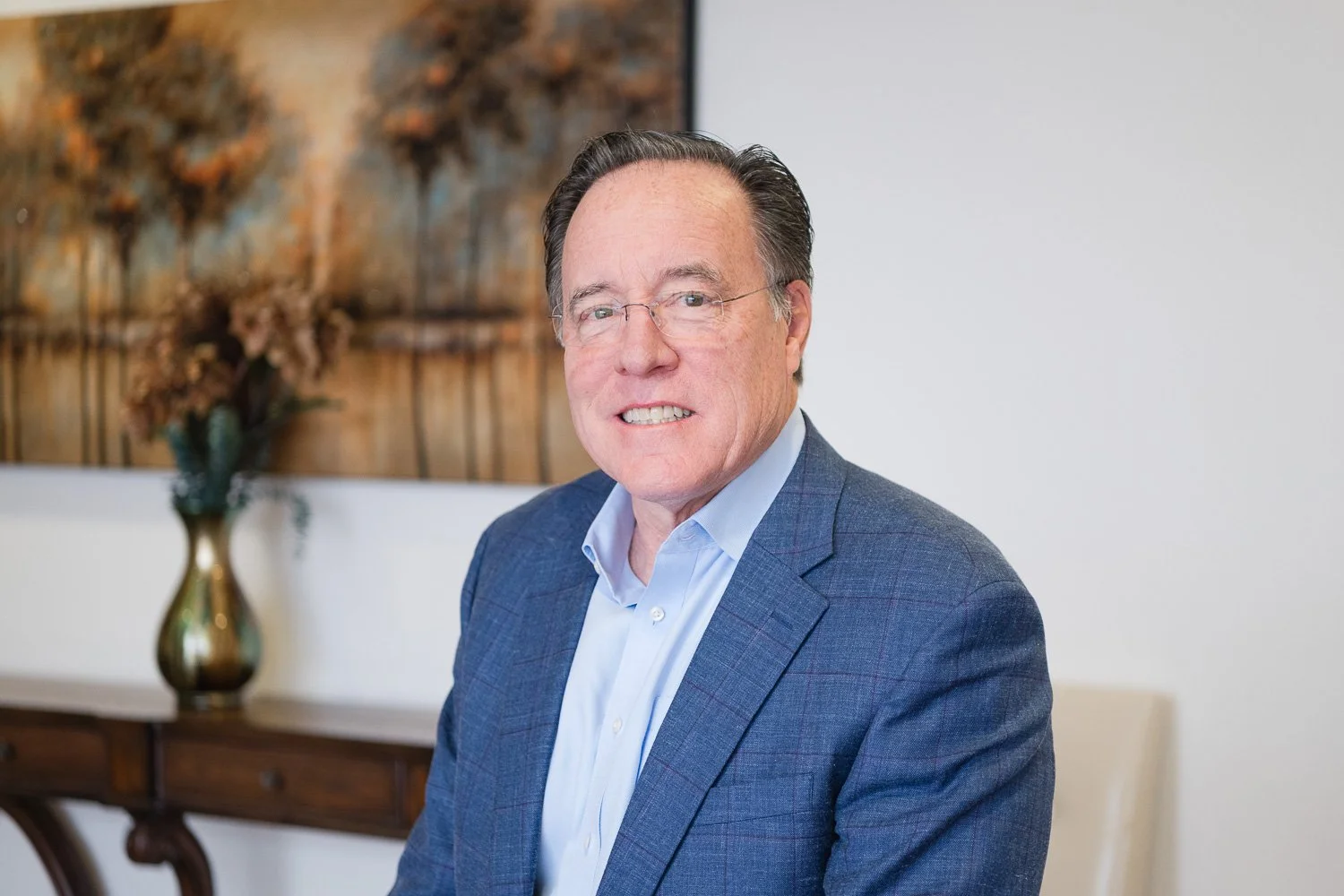 A middle-aged man wearing glasses, a blue suit jacket, and a light blue shirt, sitting in a room with a wooden sideboard and a decorative vase with dried flowers in the background.