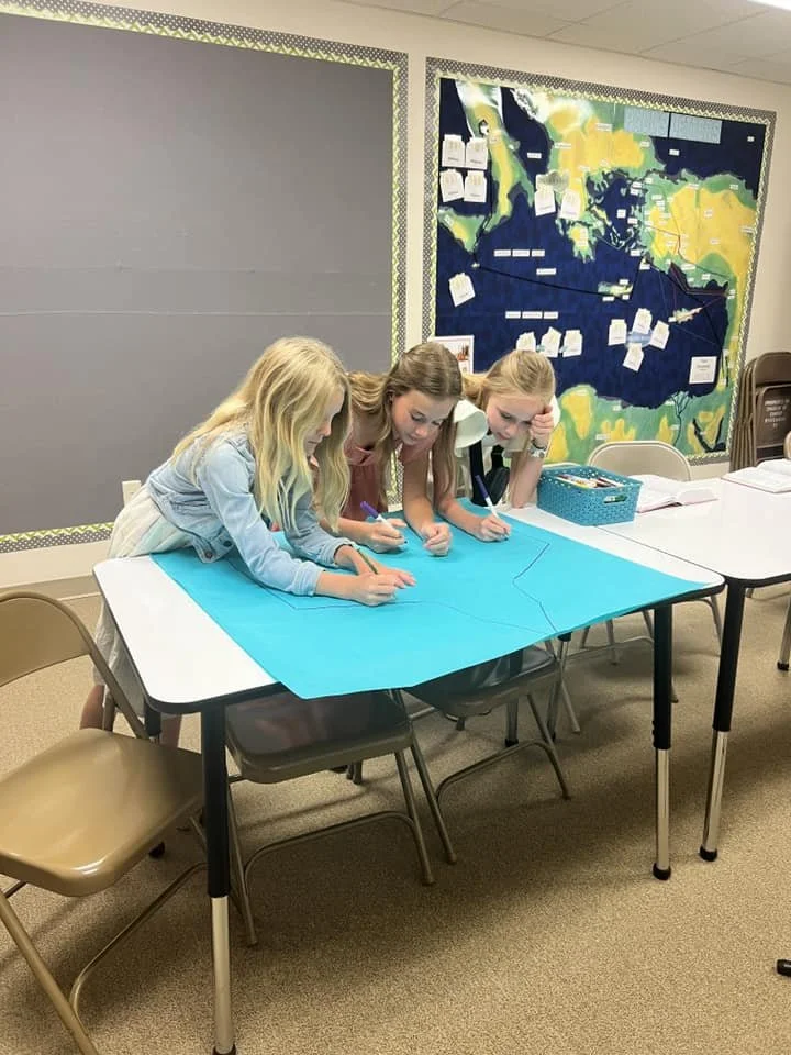 Three young girls are working together on a large blue map or poster on a table in a bible classroom. The classroom has a bulletin board with a large, colorful map and notes or papers pinned to it in the background.