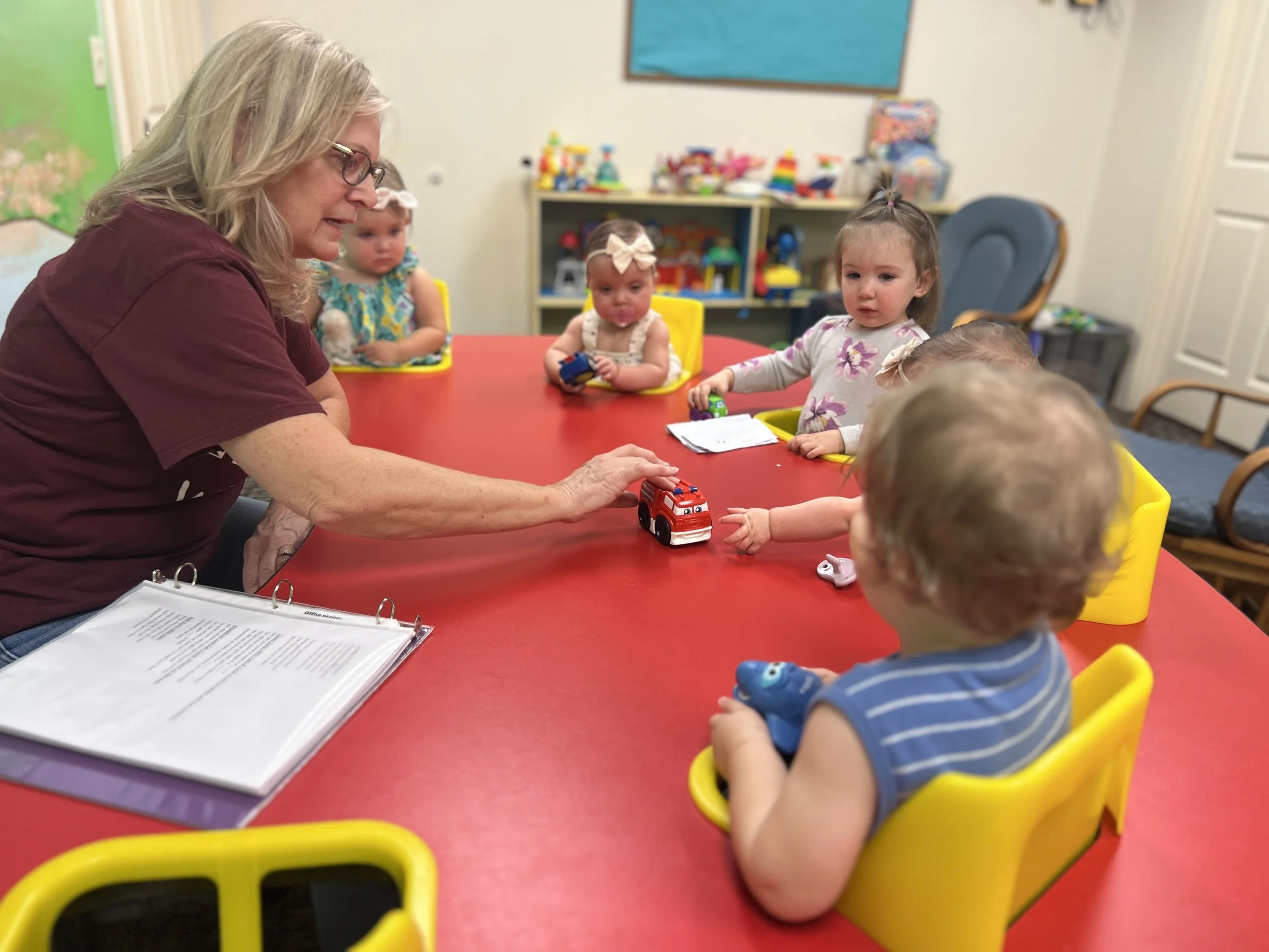 A woman interacts with five young children seated at a red table in a classroom. The woman is reaching toward a toy fire truck, and the children are holding various toys. The children have tray tables in front of them and are in yellow high chairs. T