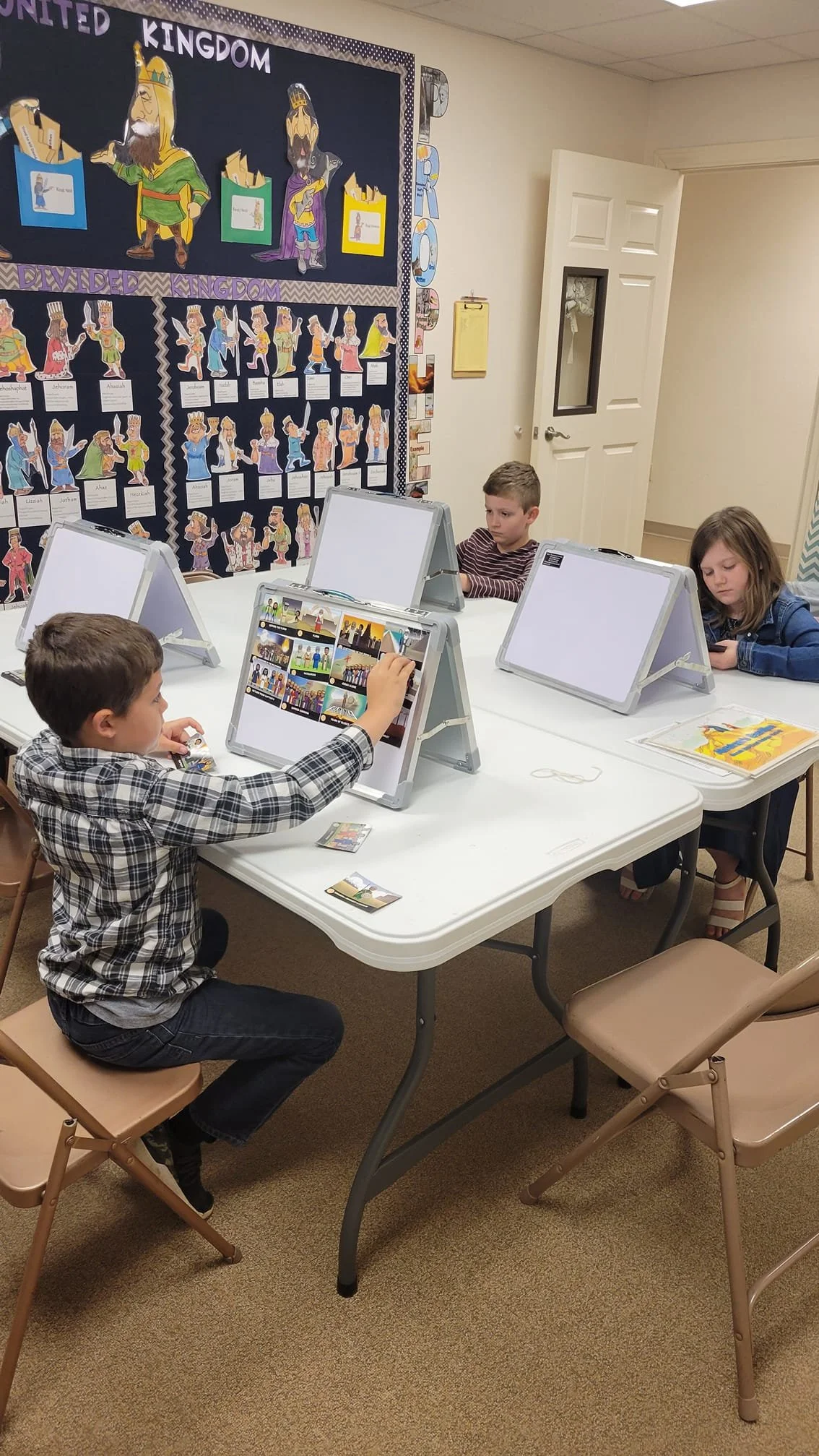 Children sitting at a table using tablets in a classroom with a biblical kingdom-themed bulletin board on the wall.