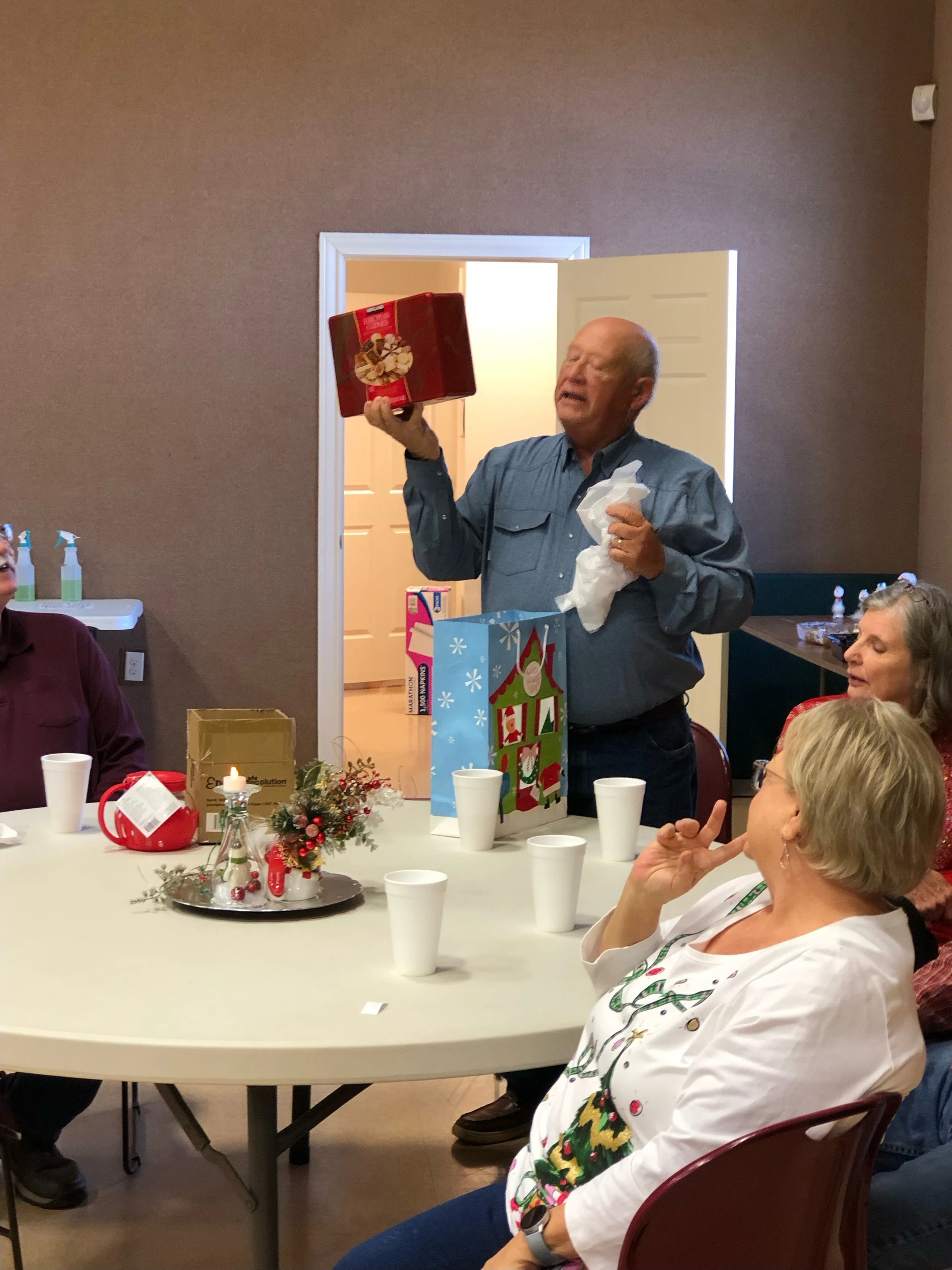 A man standing at a table, holding a gift box in one hand and a tissue in the other, opening presents during a holiday gathering. Several people sit around the table, which is decorated with holiday ornaments, candles, and gift bags.