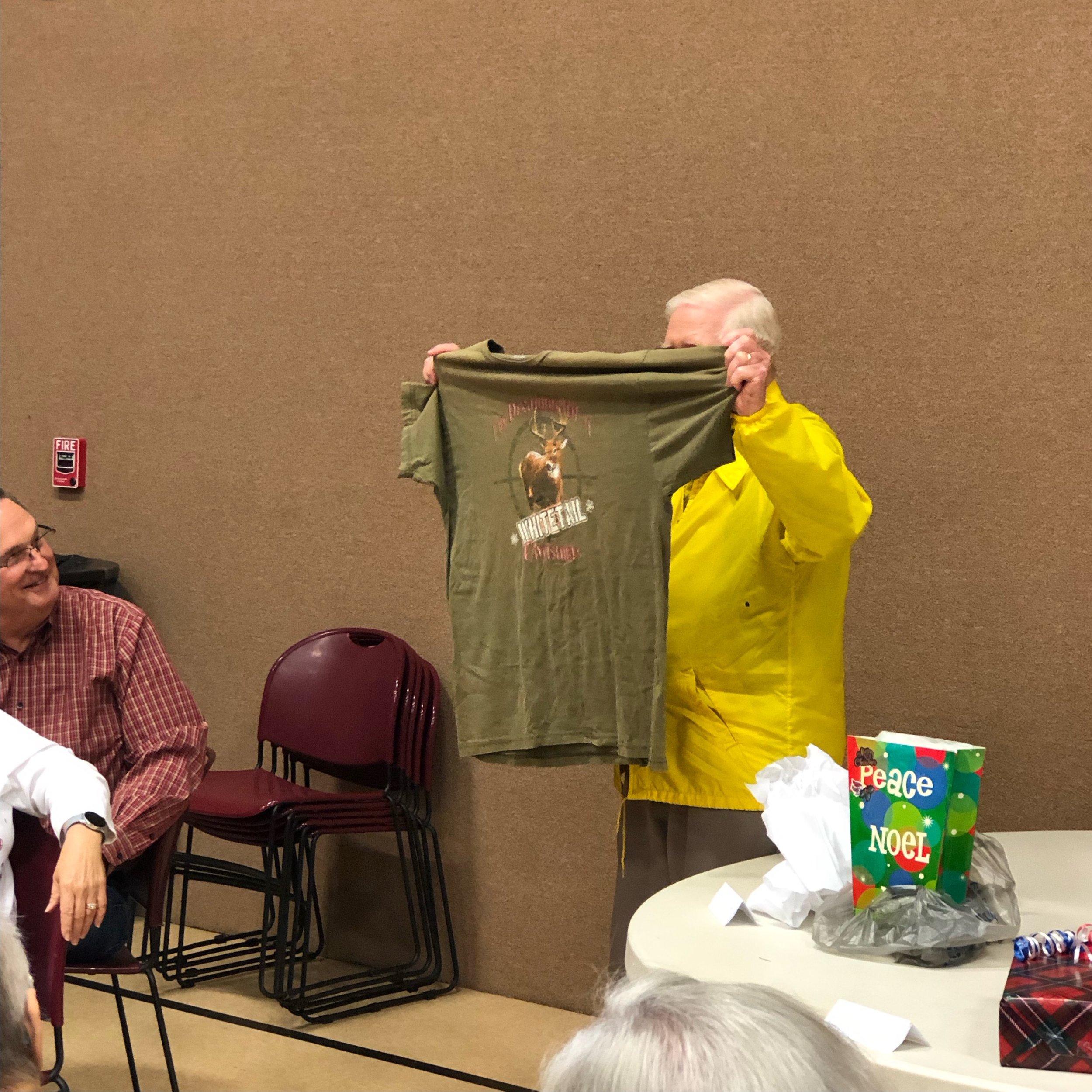 An elderly man with white hair in a yellow jacket holding up a green t-shirt with a deer and text design at a social gathering, with a smiling man in glasses and a red checkered shirt seated nearby and a table with holiday gifts in front of them.