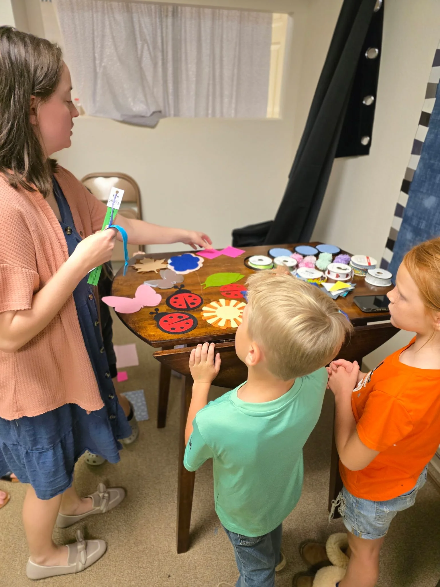 Three children at a table looking at colorful felt animal and insect cutouts, with more cutouts and craft supplies on the table.