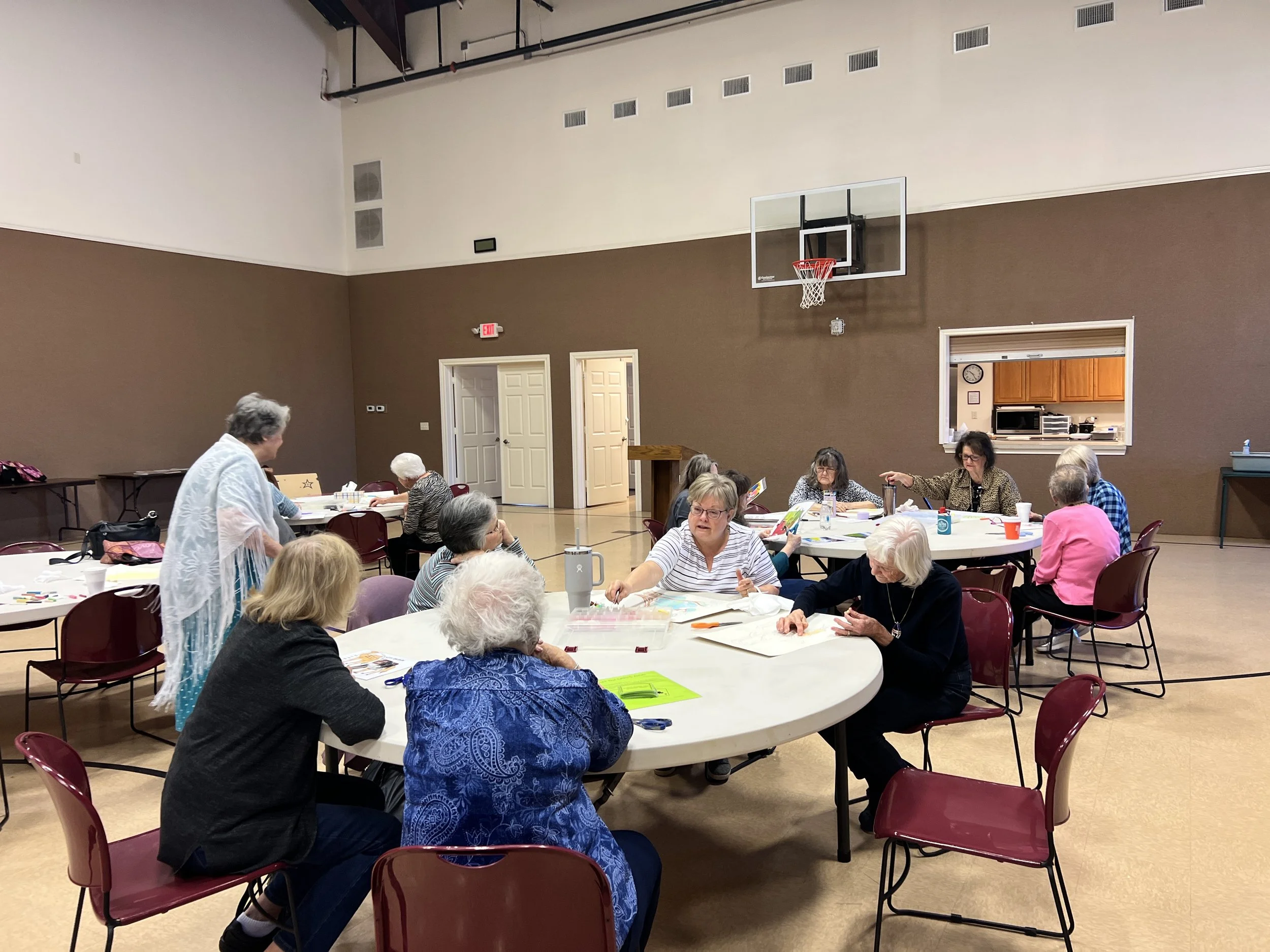Group of elderly women sitting around circular tables in a community room, engaging in arts and crafts activities.