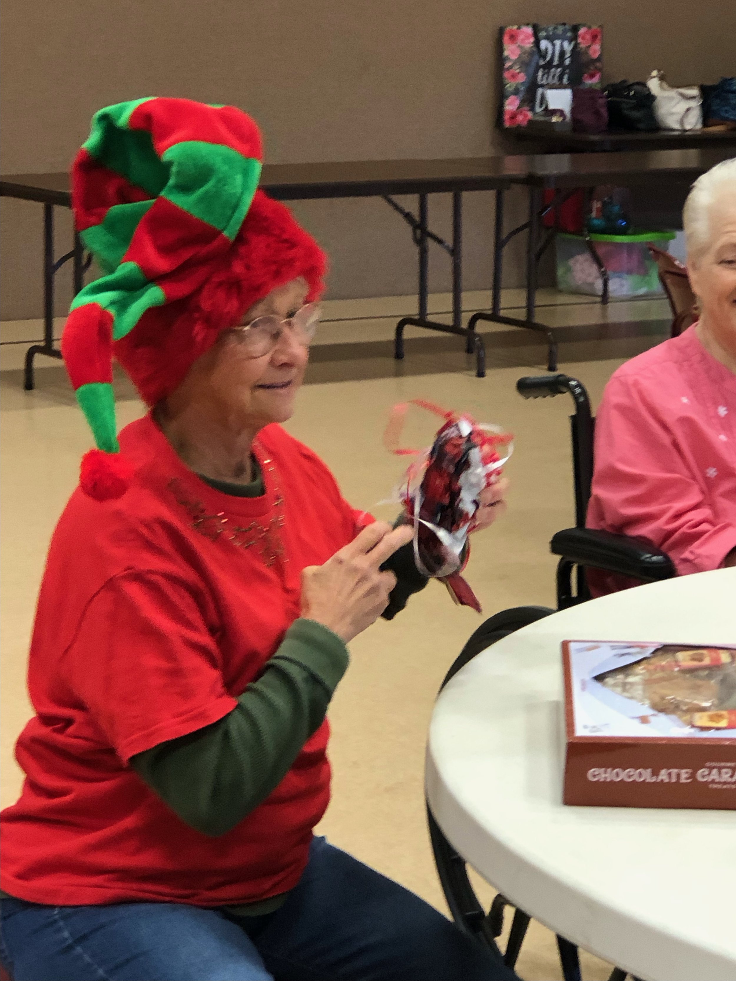 Elderly woman wearing a green and red elf hat, a red shirt, and a long sleeve green shirt underneath, sitting at a table with a box of chocolates in front of her, opening a small Christmas gift, at a holiday gathering.