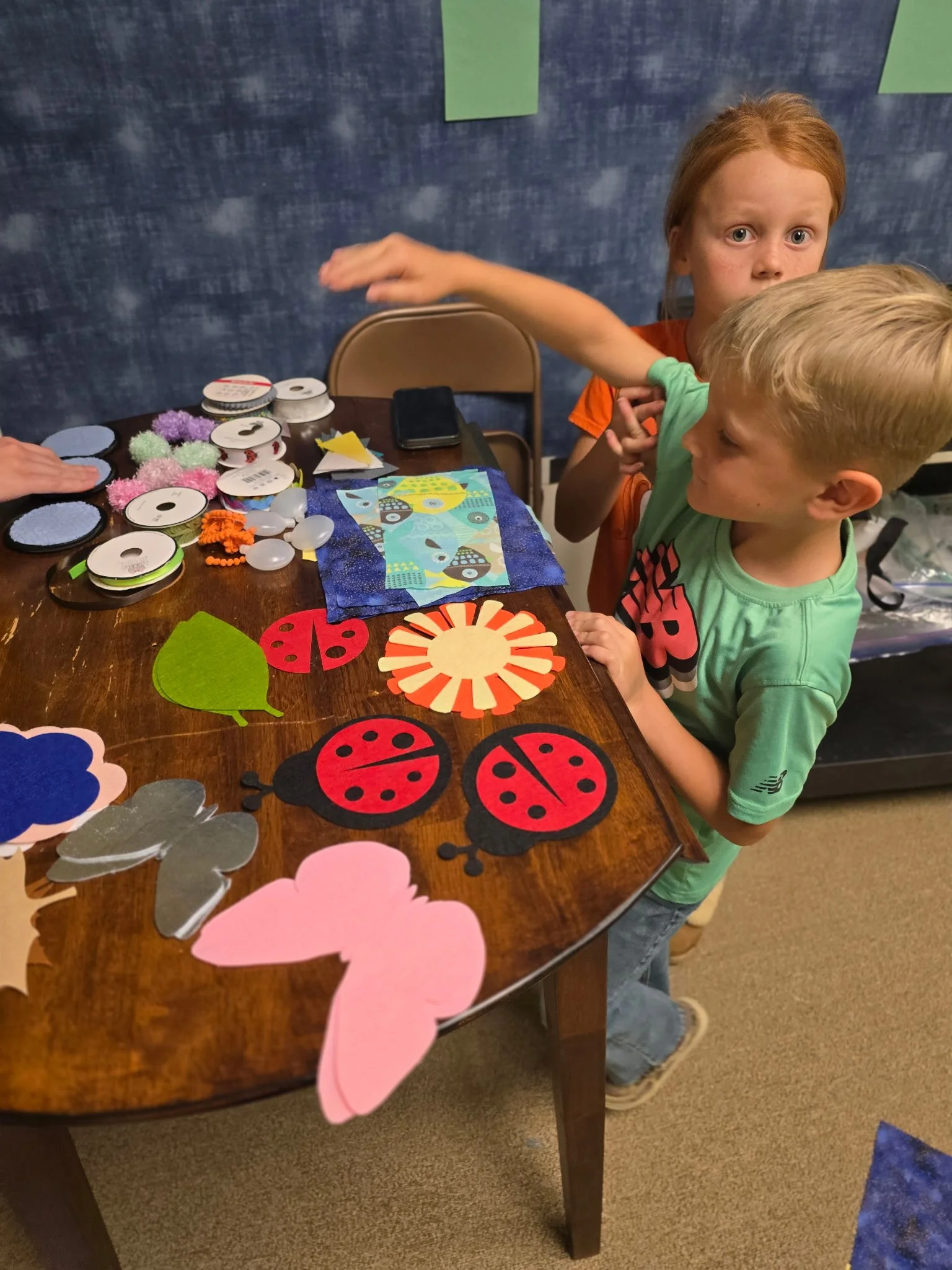 Two children at a table with various colorful craft materials, including ladybug cutouts, pom-poms, and fabric pieces for a craft project.