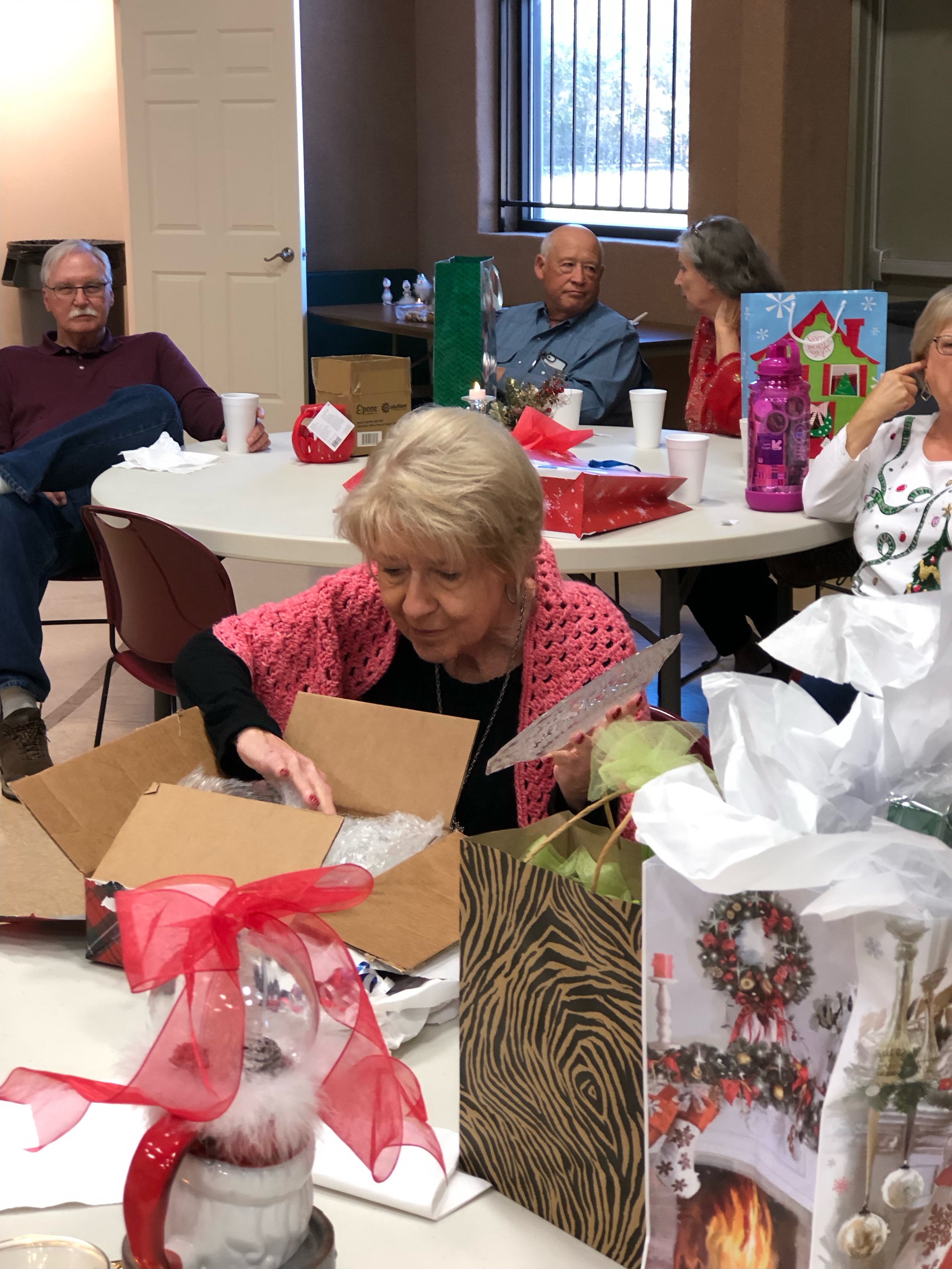 People gathered around a table with Christmas gifts and decorations, women opening presents, and others sitting and talking.
