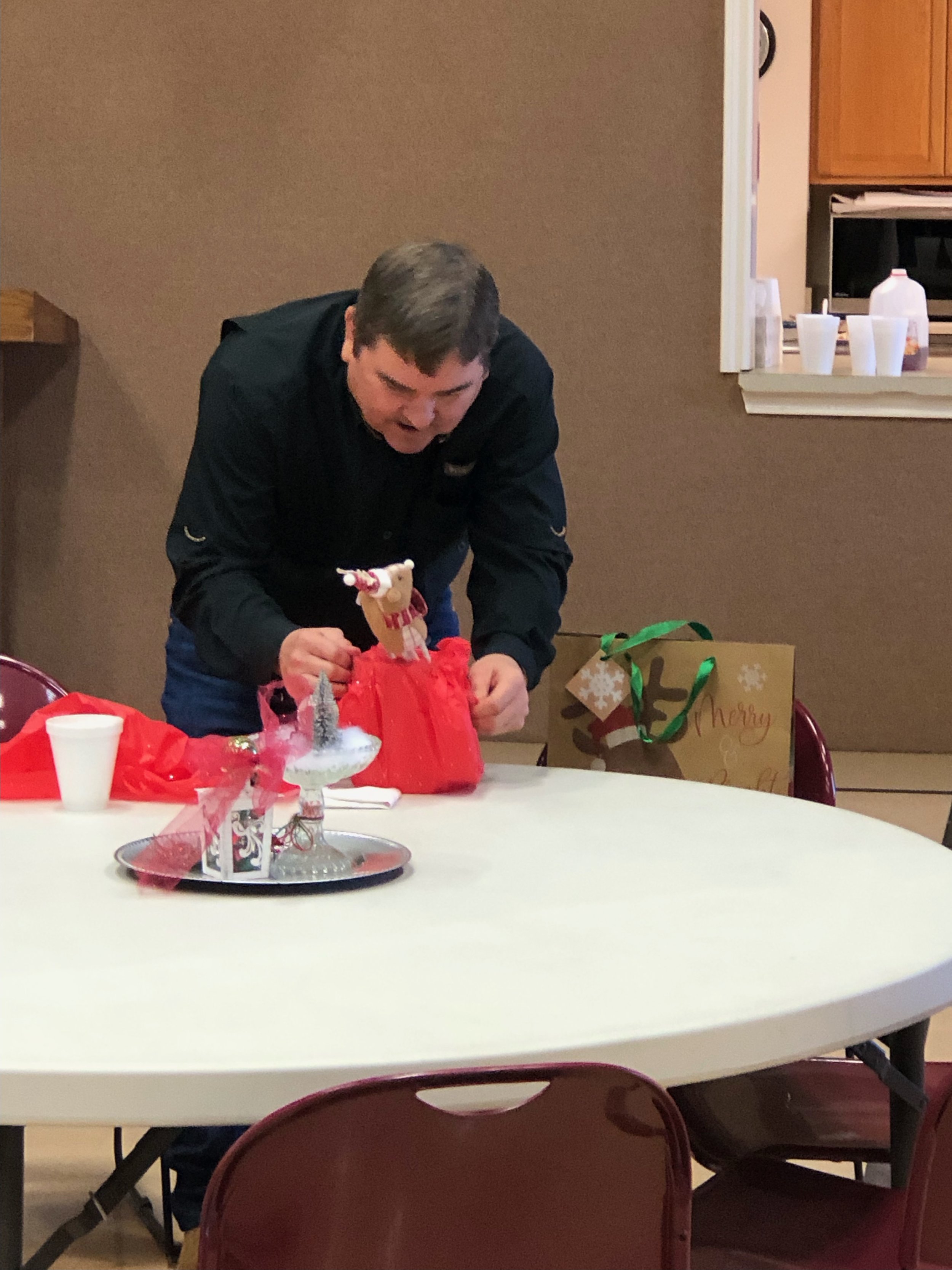 A man in black shirt leaning over a table opening a red gift bag, with Christmas decorations and presents on the table and behind him.