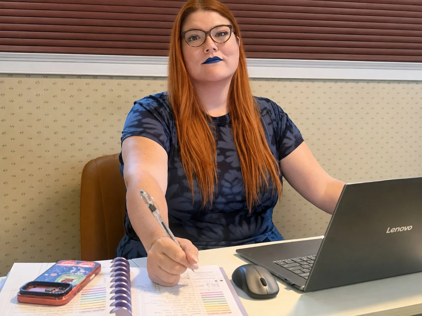 A woman with long red hair and glasses sitting at a table, writing in a planner with a pen. She has blue lipstick and is wearing a blue and black patterned shirt, with a laptop, mouse, and colorful phone case on the table.