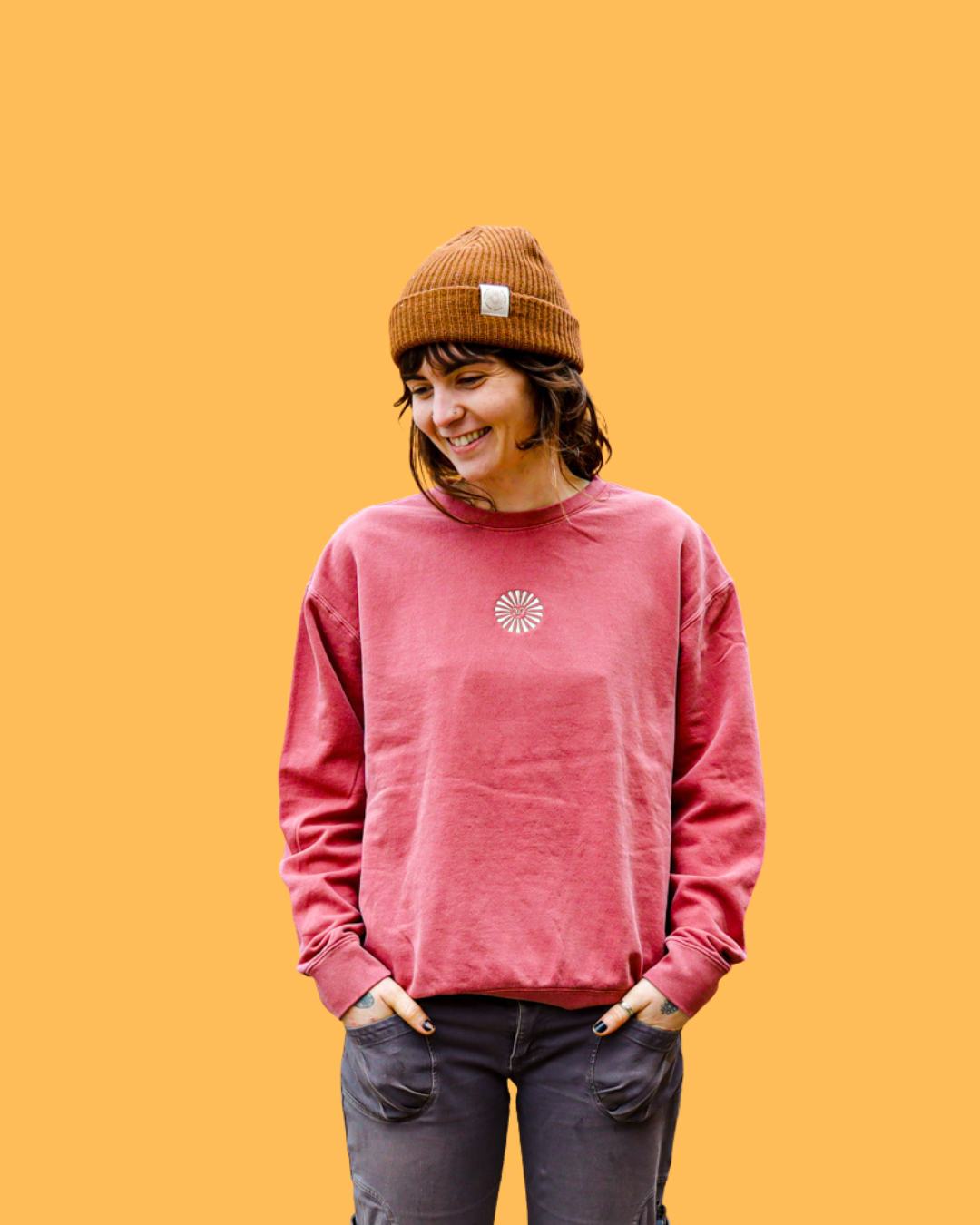 A woman with shoulder-length dark hair wearing a brown beanie, a pink Empire crewneck, and dark gray pants, smiling with her hands in her pockets, standing against a plain orange background.