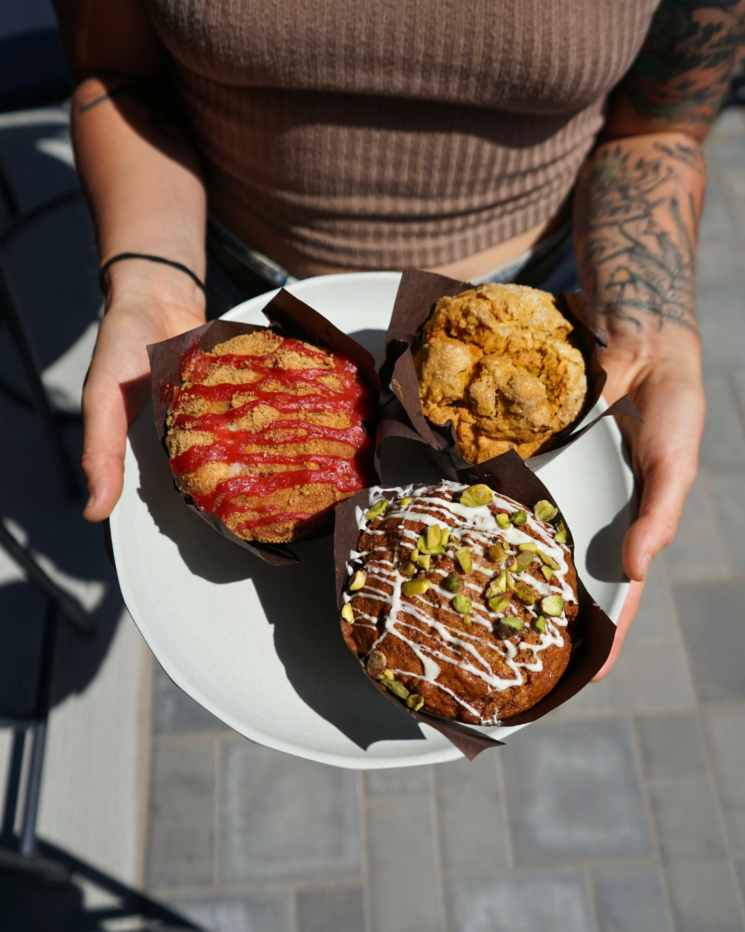 Person holding a white plate with three muffins topped with various glazes and toppings outside on a paved surface.