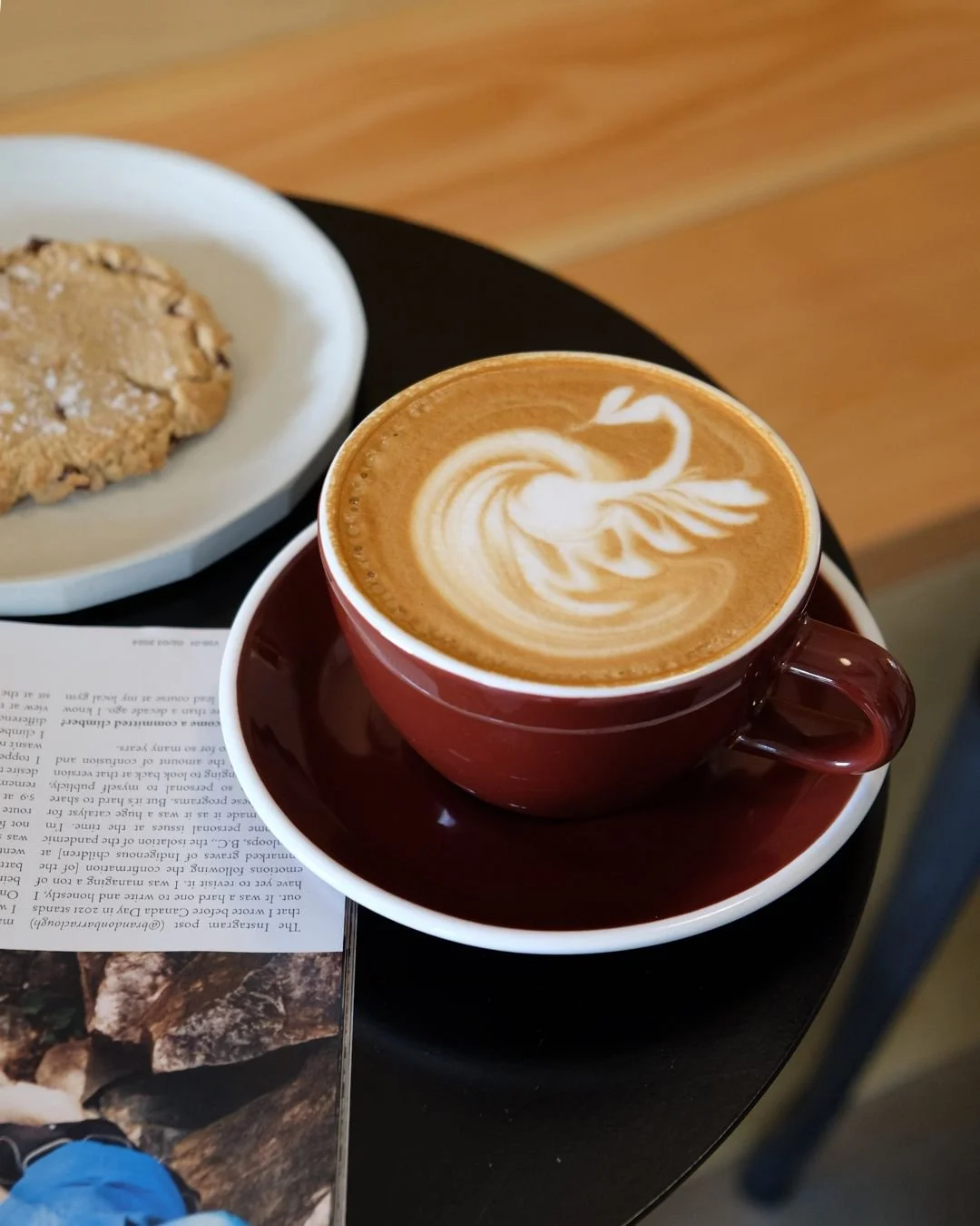 A red cup of coffee with latte art on top, sitting on a saucer. Next to it, a white plate with a large chocolate chip cookie. Part of a magazine or newspaper is visible beneath the saucer.