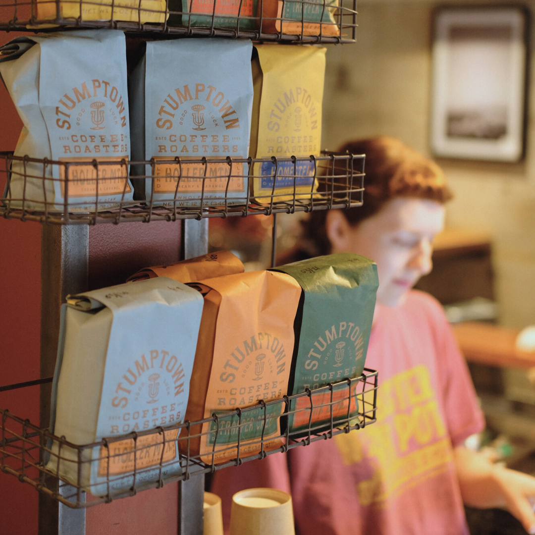 Coffee bags labeled 'Stumptown Coffee Roasters' on metal wire racks in a coffee shop, with a person in a pink shirt blurred in the background.