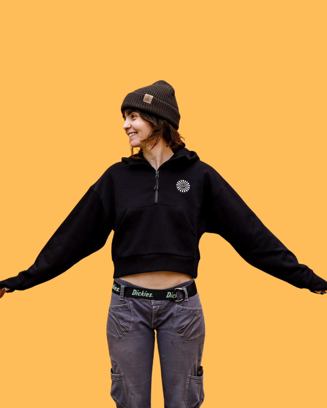 A woman smiling and looking to the side, wearing a black Empire quarter zip cropped hoodie, dark gray cargo pants, and a dark brown beanie against a solid orange background.