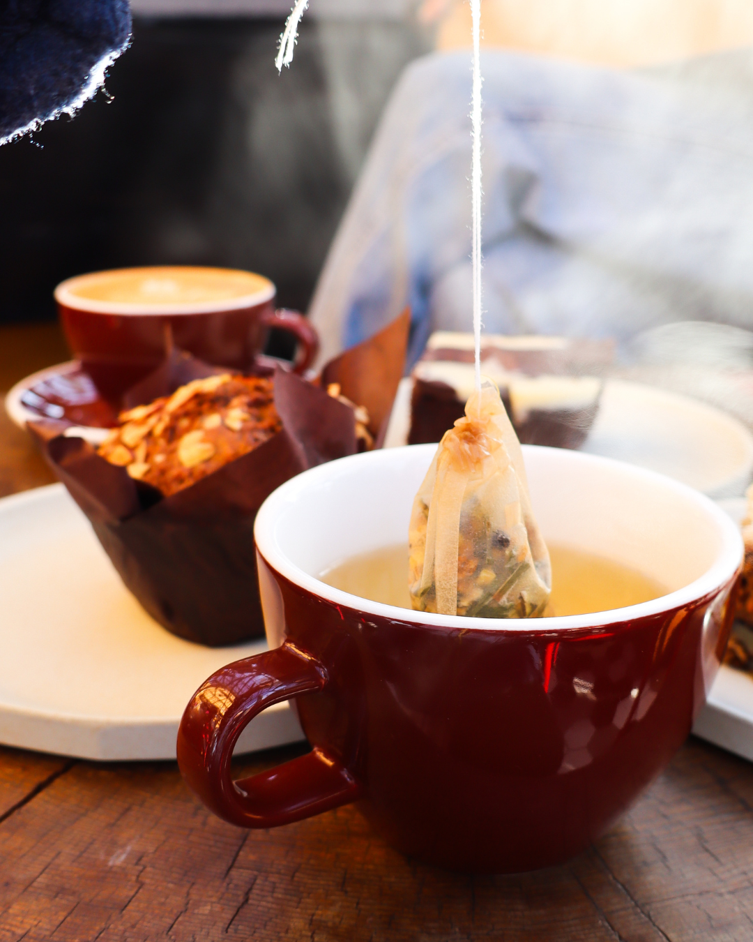 A teabag with tea steeping inside a maroon ceramic mug, with a plate with a muffin and another cup of tea in the background.
