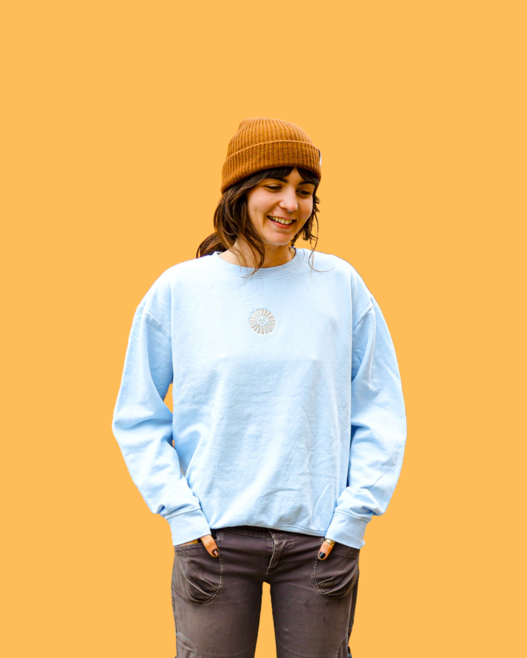 A young woman is smiling and looking down, wearing a brown beanie, a light blue Empire sweatshirt, and dark pants, against a solid orange background.