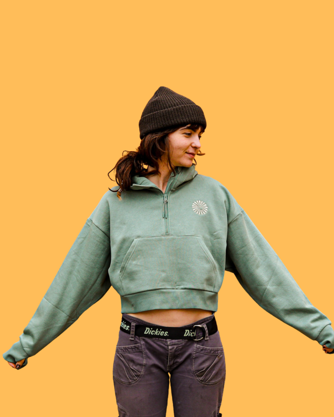 Young woman in a green Empire quarter zip cropped hoodie, and black pants, wearing a brown beanie against a yellow background.