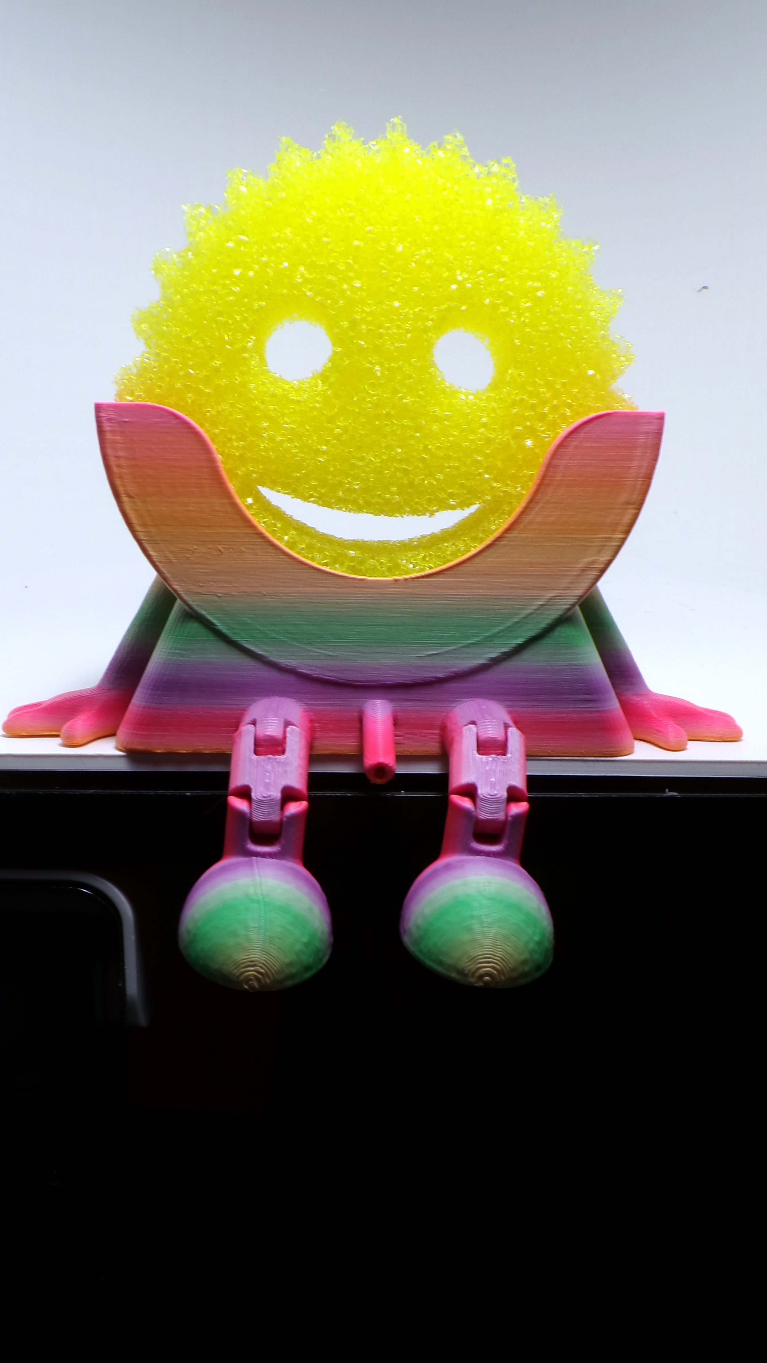 3D printed frog with a yellow smiley face made of textured material, sitting on a rainbow-colored base with pink webbed feet.