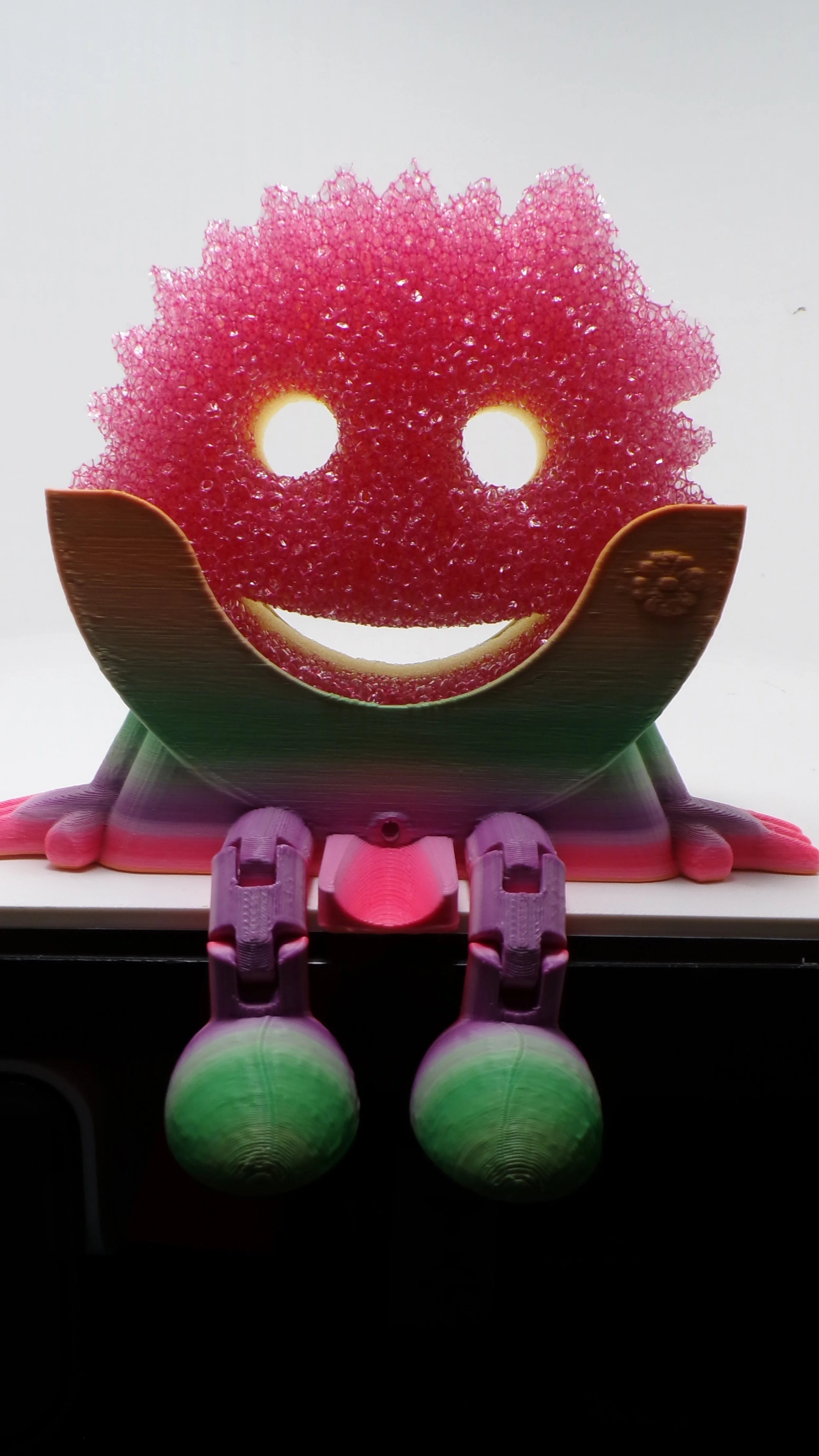 A cellphone case designed as a frog with a smiling face, with the face made from pink textured material and the body from pink and green parts.