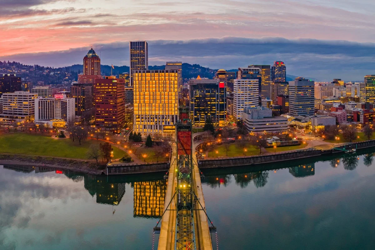 An aerial view of Portland, Oregon skyline during dusk, showing tall buildings and a river with reflections of the city lights.