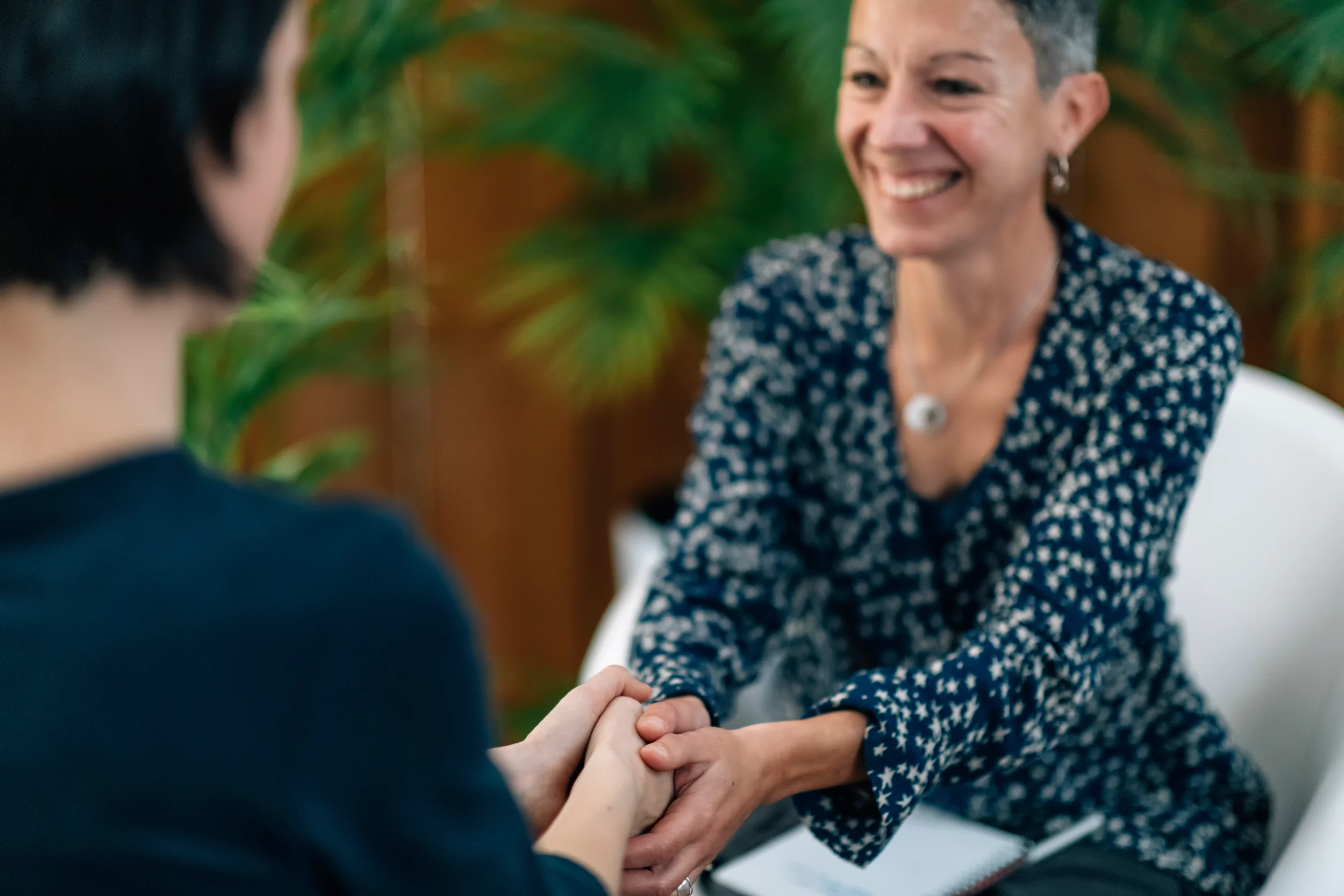 A woman smiling and holding the hands of another person, sitting in a room with green plants and wooden furniture.