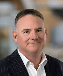 Professional man with short grey hair and in a dark pinstripe suit, smiling, in a blurred office background.