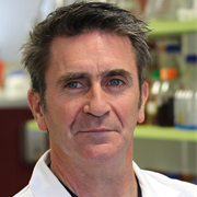 A man with dark hair and blue eyes, wearing a white lab coat, standing in a laboratory with shelves of scientific equipment and supplies in the background.