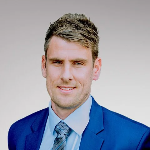 A man with short brown hair wearing a blue suit, light blue shirt, and striped tie, smiling at the camera against a plain gray background.