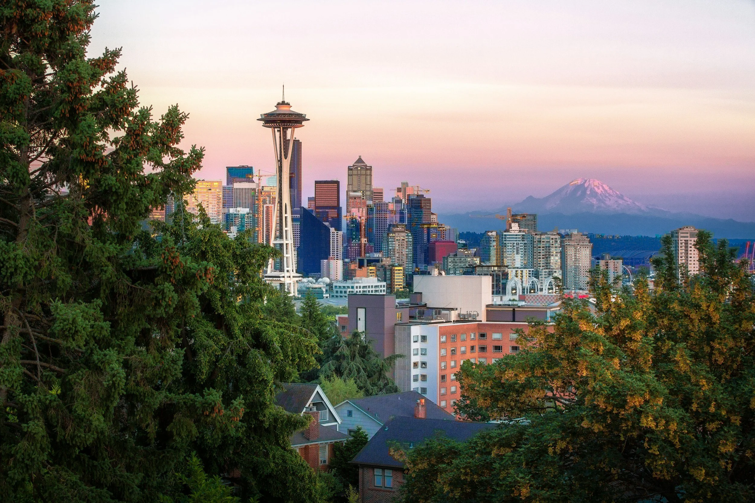 View of downtown Seattle with the Space Needle, high-rise buildings, trees in the foreground, and Mount Rainier in the background at sunset.
