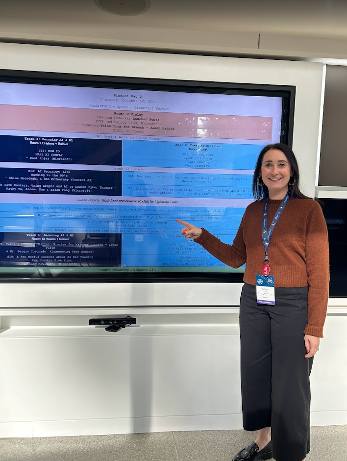 A woman with brown hair and a orange sweater pointing at a digital conference schedule on a large screen, with a lanyard and conference badge, in a modern conference room.