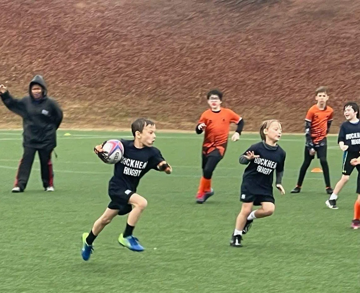 Young children playing rugby on a field, with some running and others standing, some wearing black jerseys with 'Buckhead Rugby' written on them, and a coach or adult in black in the background.