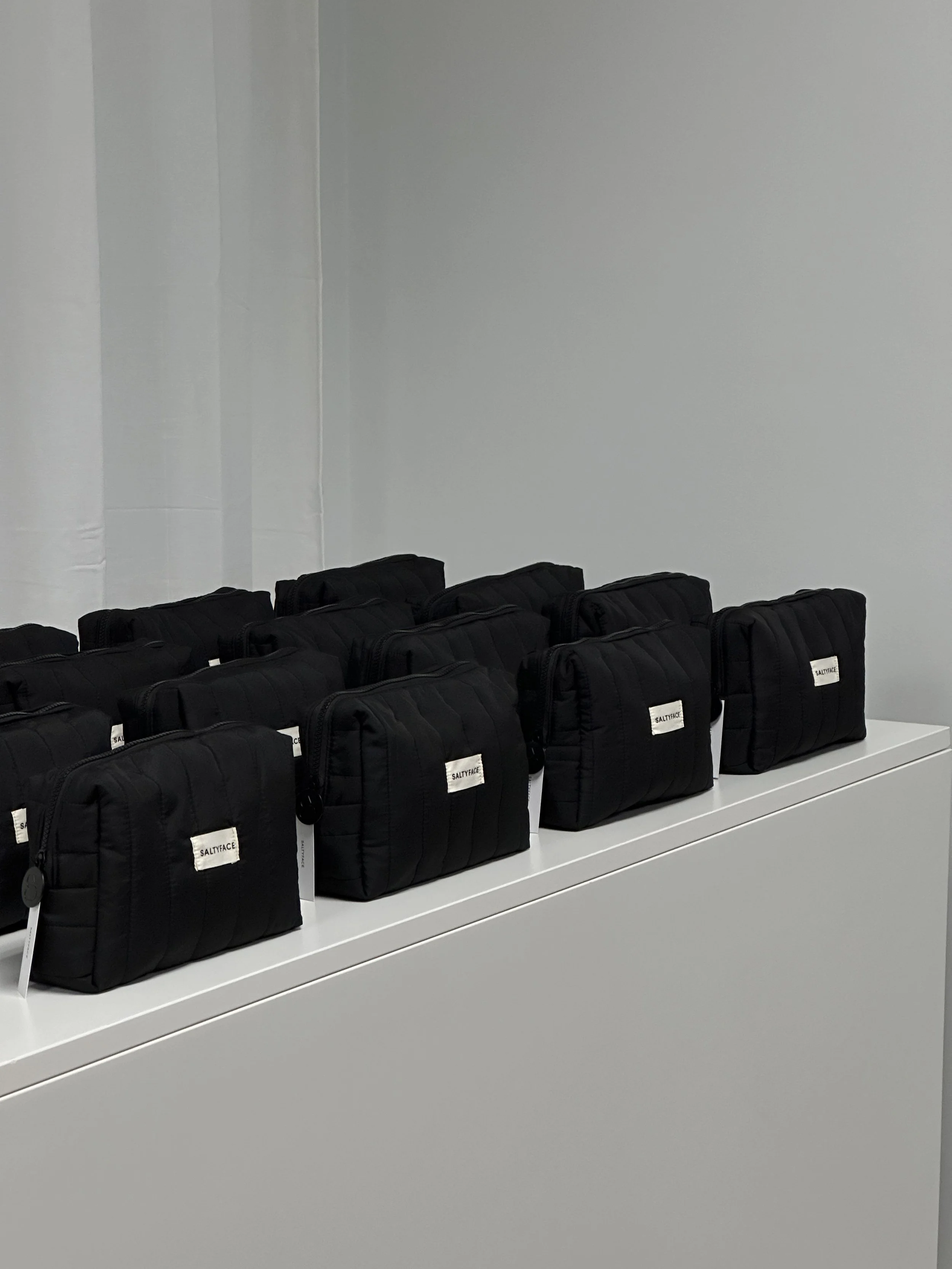 Multiple black SALTYFACE toiletry bags arranged on a white table against a light gray wall.