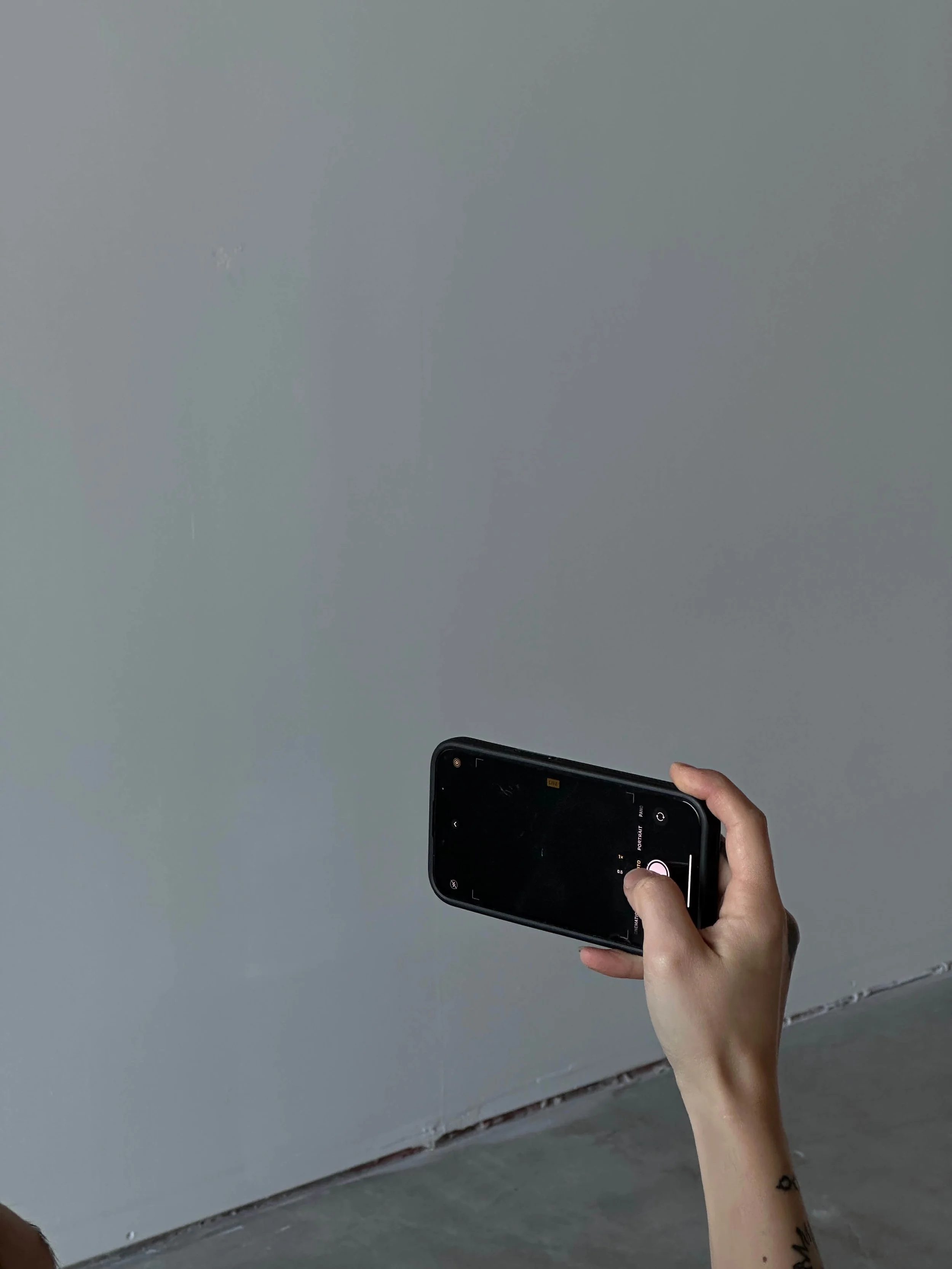 A person holding a smartphone with a black case, positioned horizontally, with a tattoo visible on their wrist, against a plain grey wall.