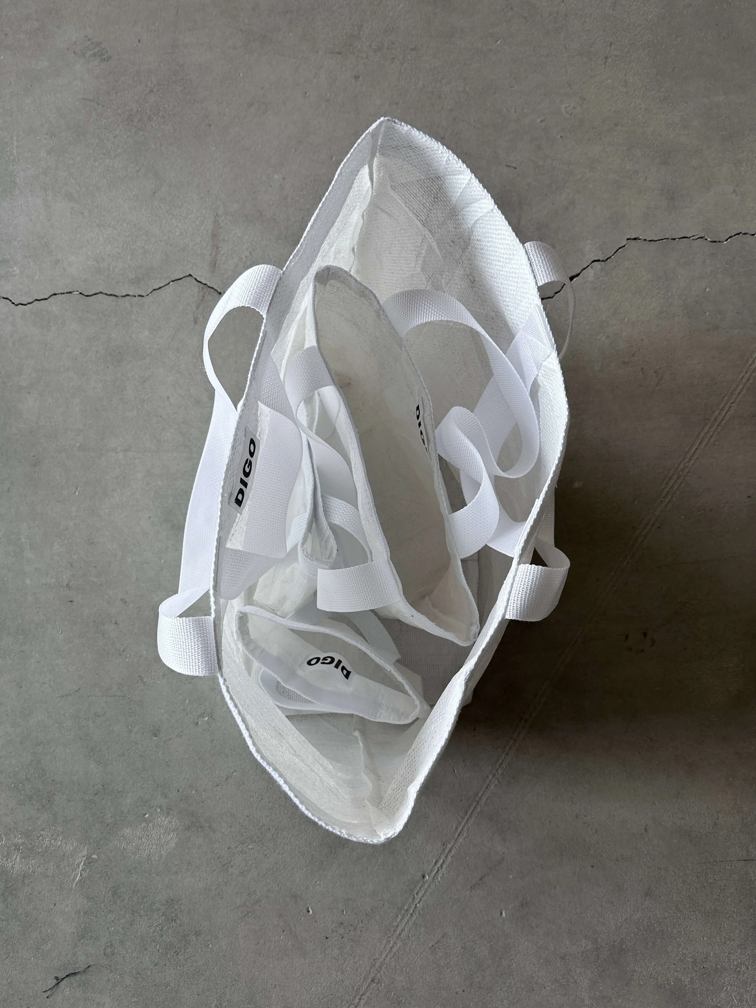 A transparent plastic bag with white handles and interior compartments, lying on a concrete floor.