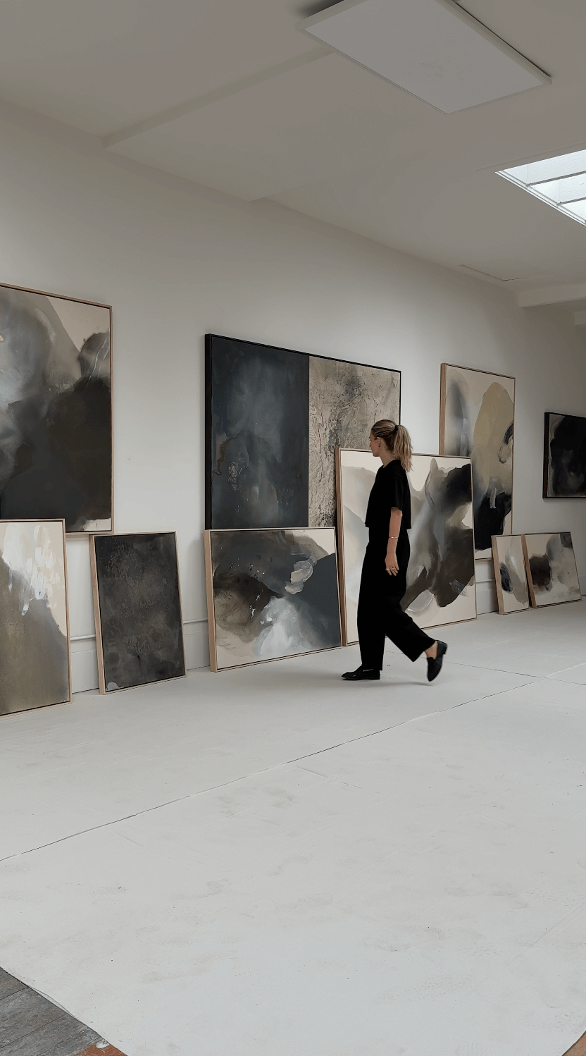 A woman walking in an art gallery with various abstract paintings displayed on the walls and leaning on the floor.