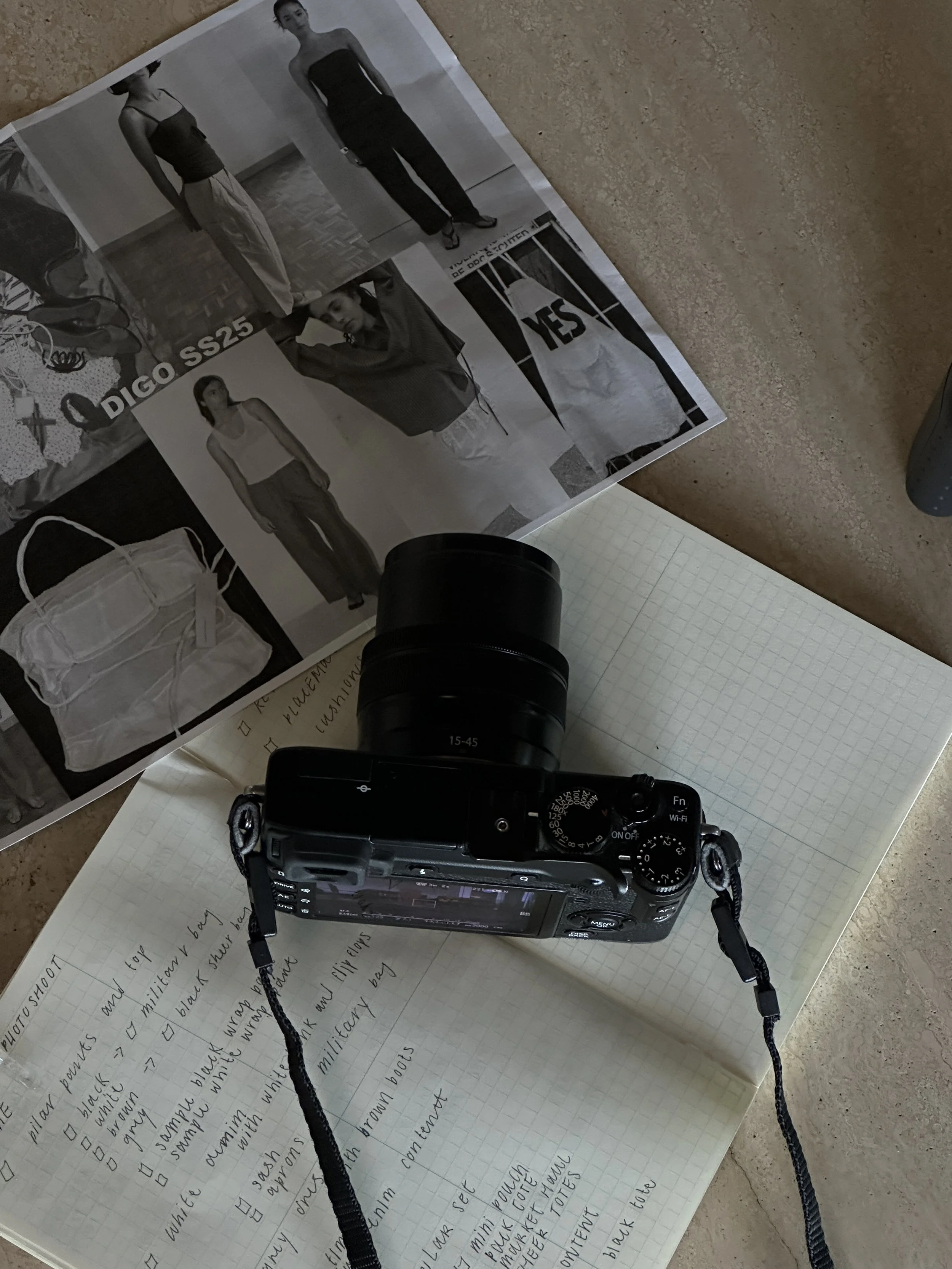 A camera resting on a grid-pattern notebook with notes, a fashion magazine for DIGO SS25 collection, and a gray carpeted surface.