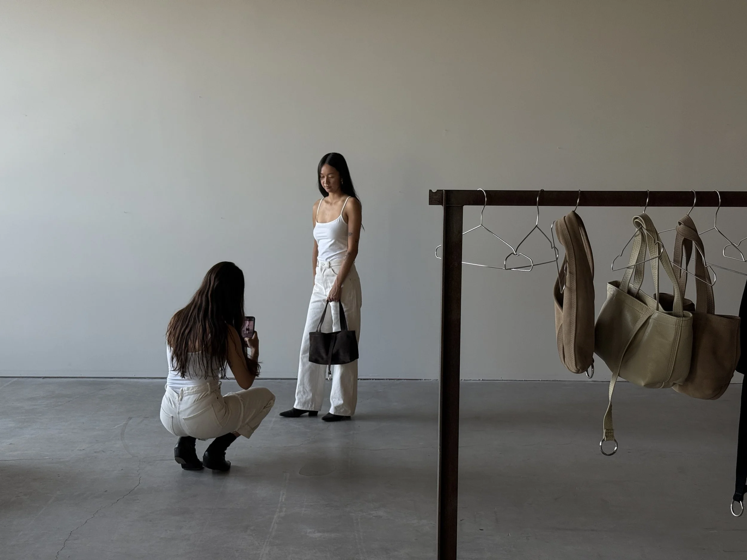 Two women in white outfits, one standing and one crouching, in a minimalistic space with a clothing rack holding beige and brown bags.