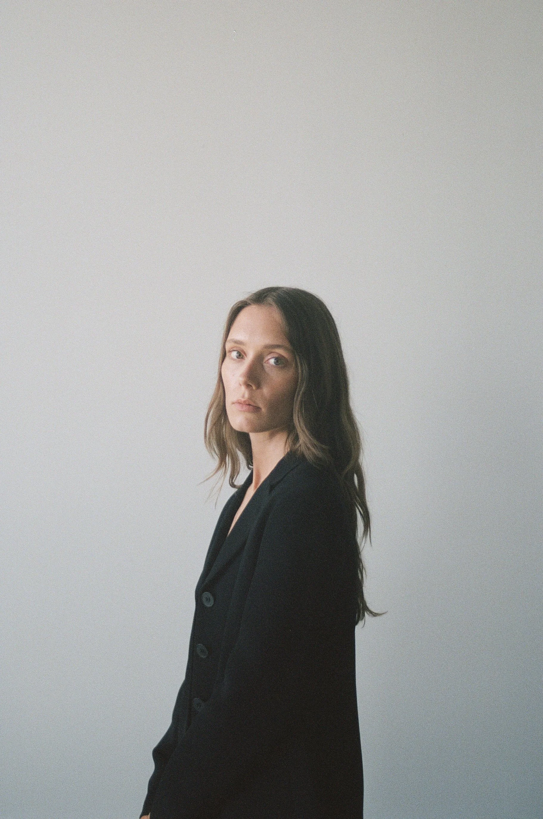 Ellie West with long wavy brown hair wearing a black blazer stands against a plain light gray background.