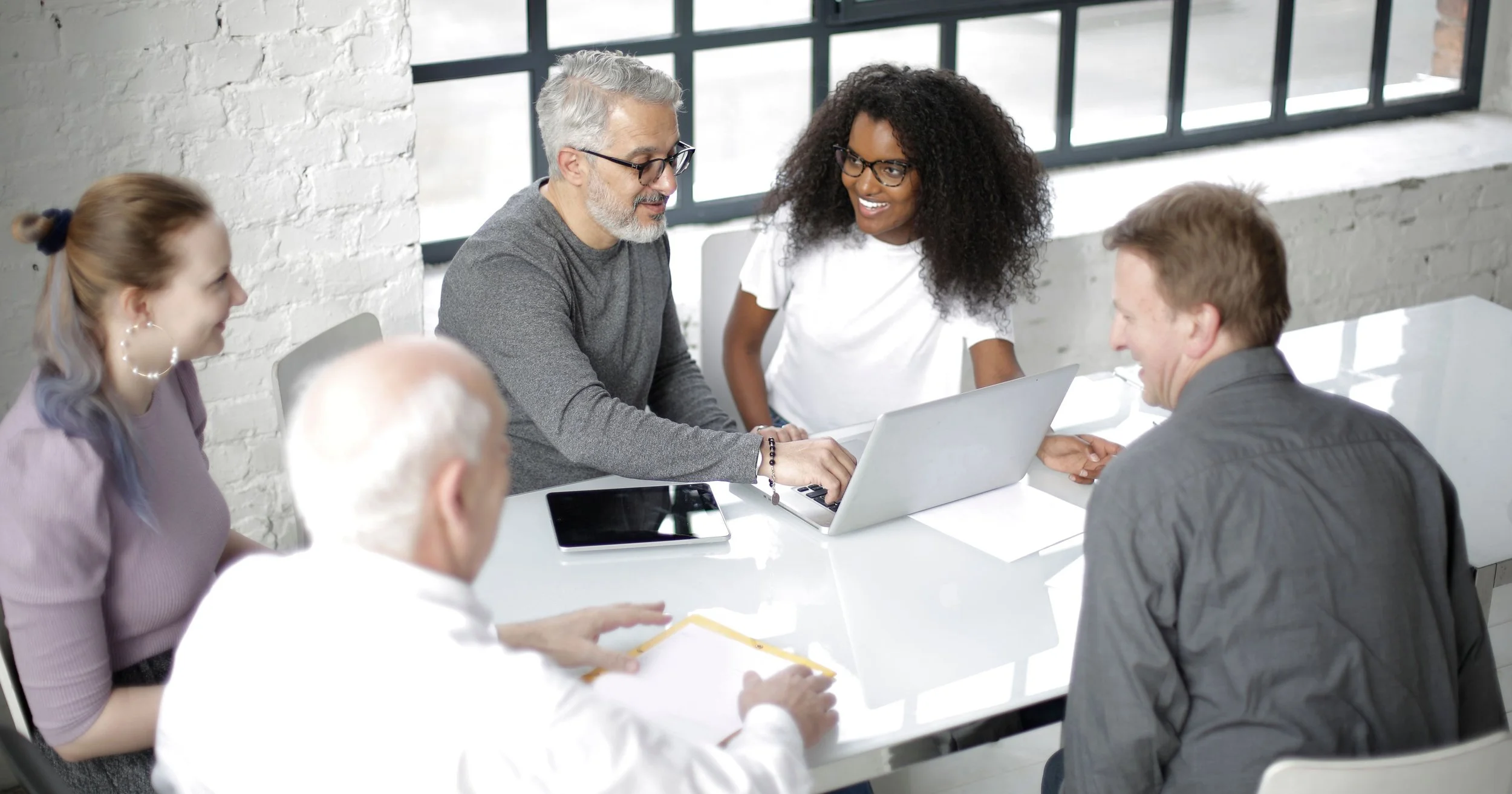 A group of professionals sit around a white table in a bright office, collaborating on a laptop while discussing work in a modern workspace.