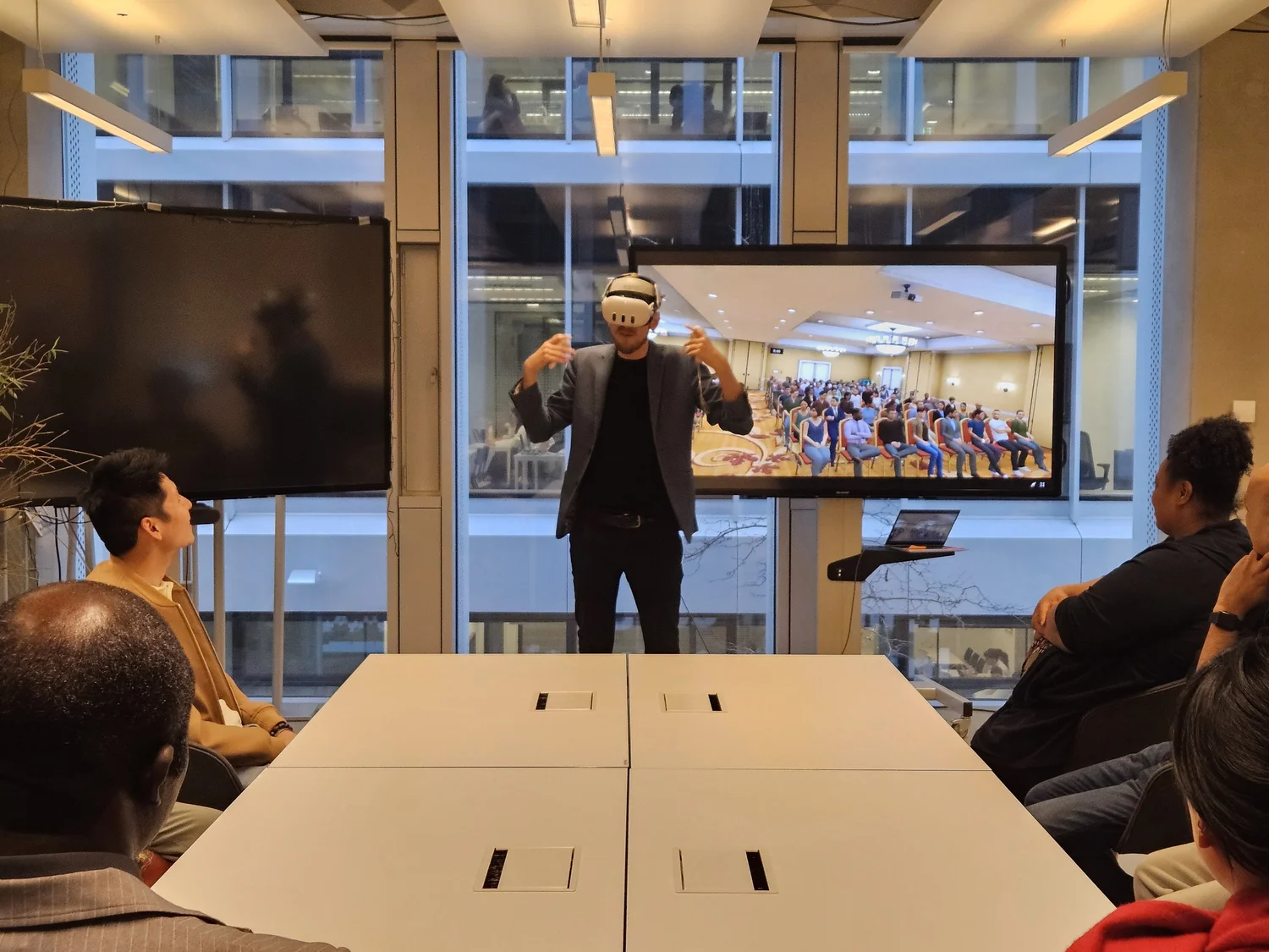 A participant in a VR headset speaks to a virtual audience shown on screen while the group watches
