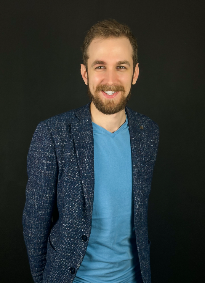 A smiling man with a beard and mustache, wearing a blue t-shirt and a dark blue blazer standing against a black background.