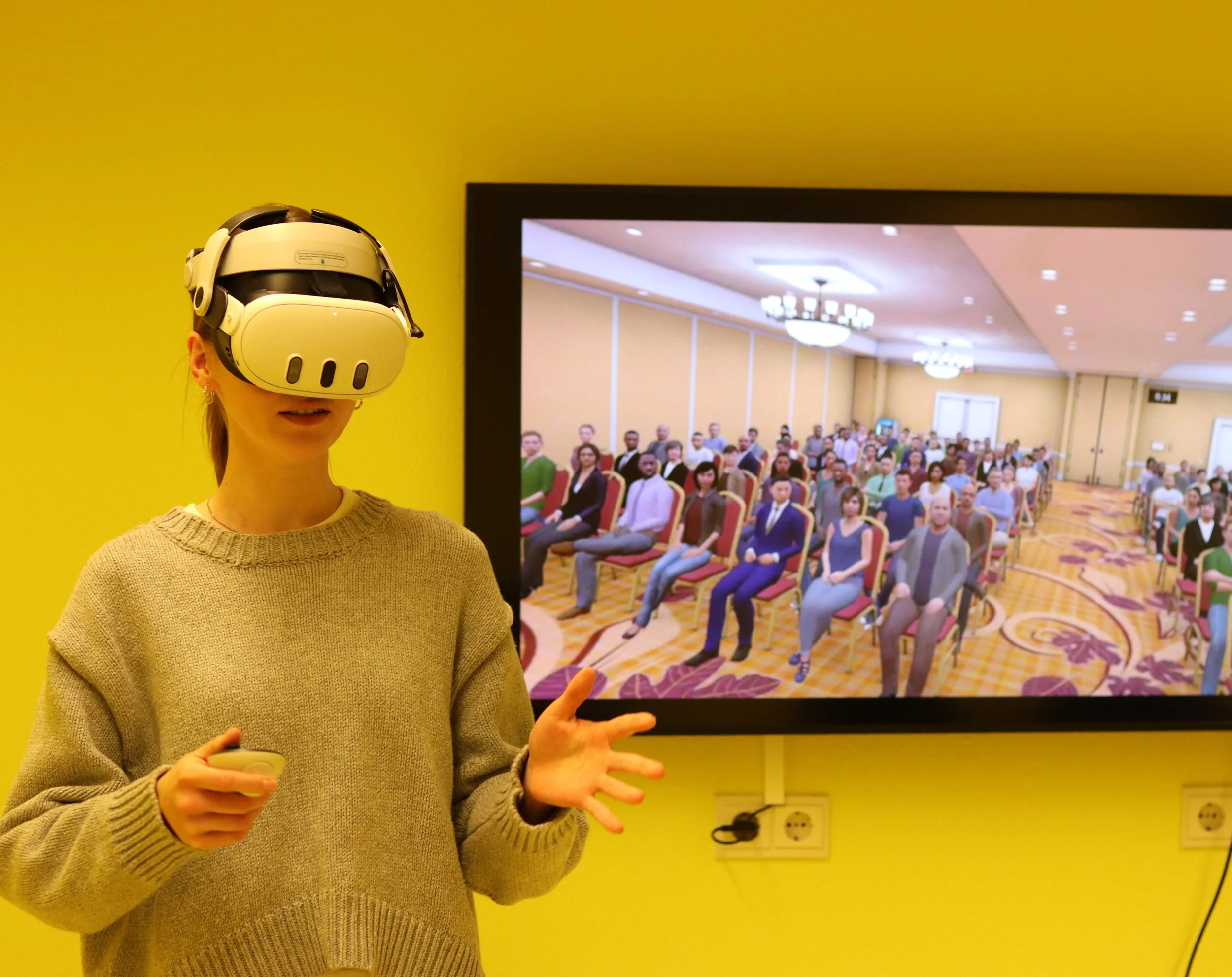 A woman wearing a virtual reality headset and holding a remote controller, standing in front of a large screen displaying a conference room filled with seated attendees.