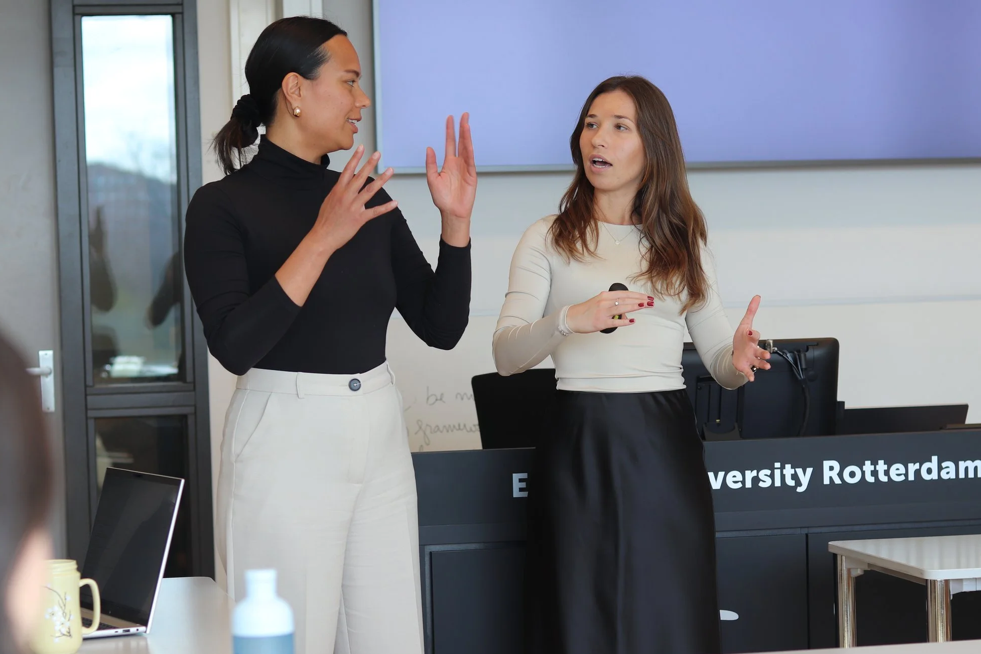Two women engaged in a discussion in a classroom or conference room. One woman is wearing a black top and white pants, and the other is wearing a white top and black skirt. There is a large screen behind them, and a computer monitor and some beverages on a table in the foreground.