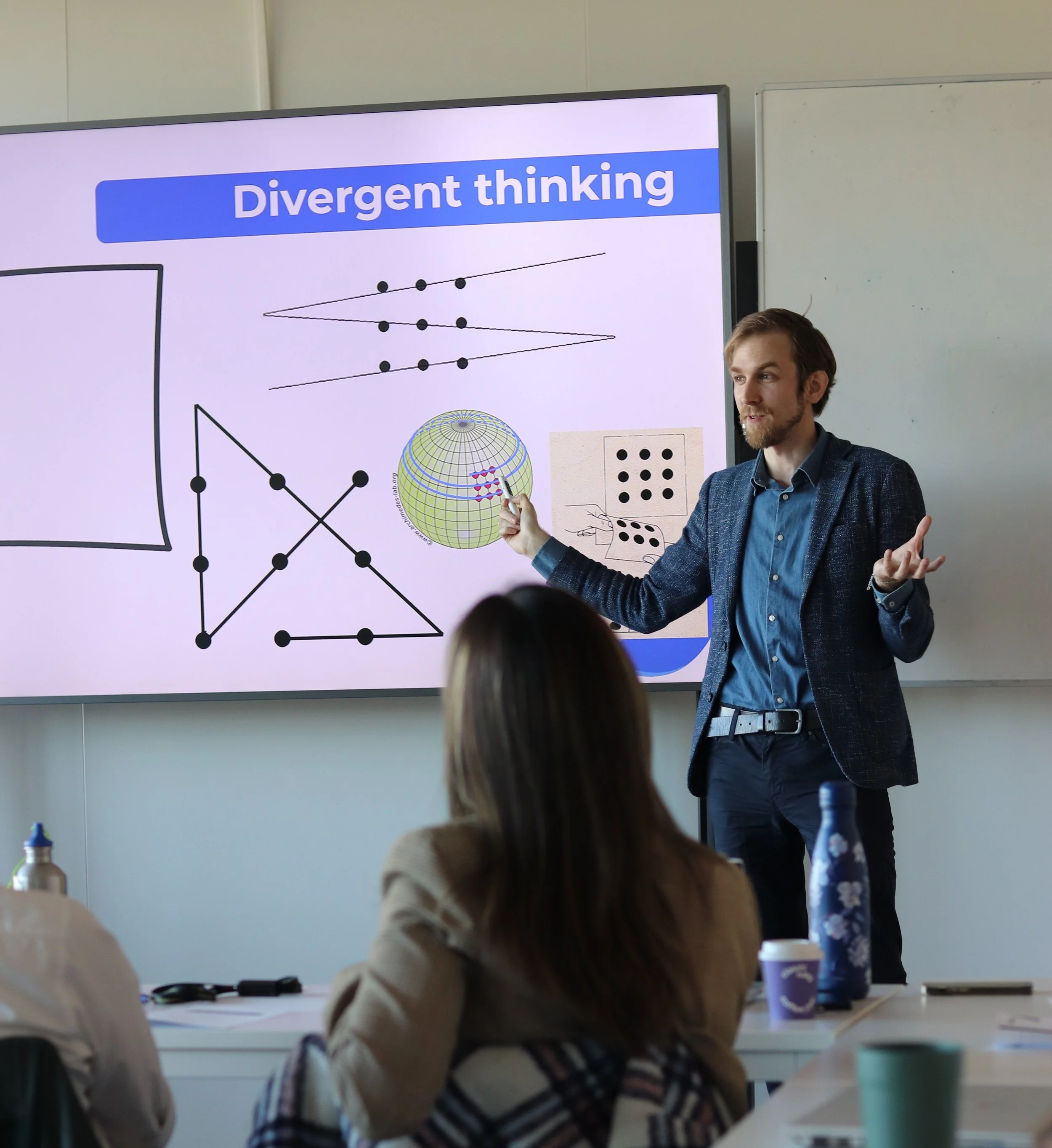 A man giving a presentation on a topic called 'Divergent thinking' in a classroom or conference room. He is standing in front of a large screen with diagrams and text, gesturing with his hands while talking to an audience.