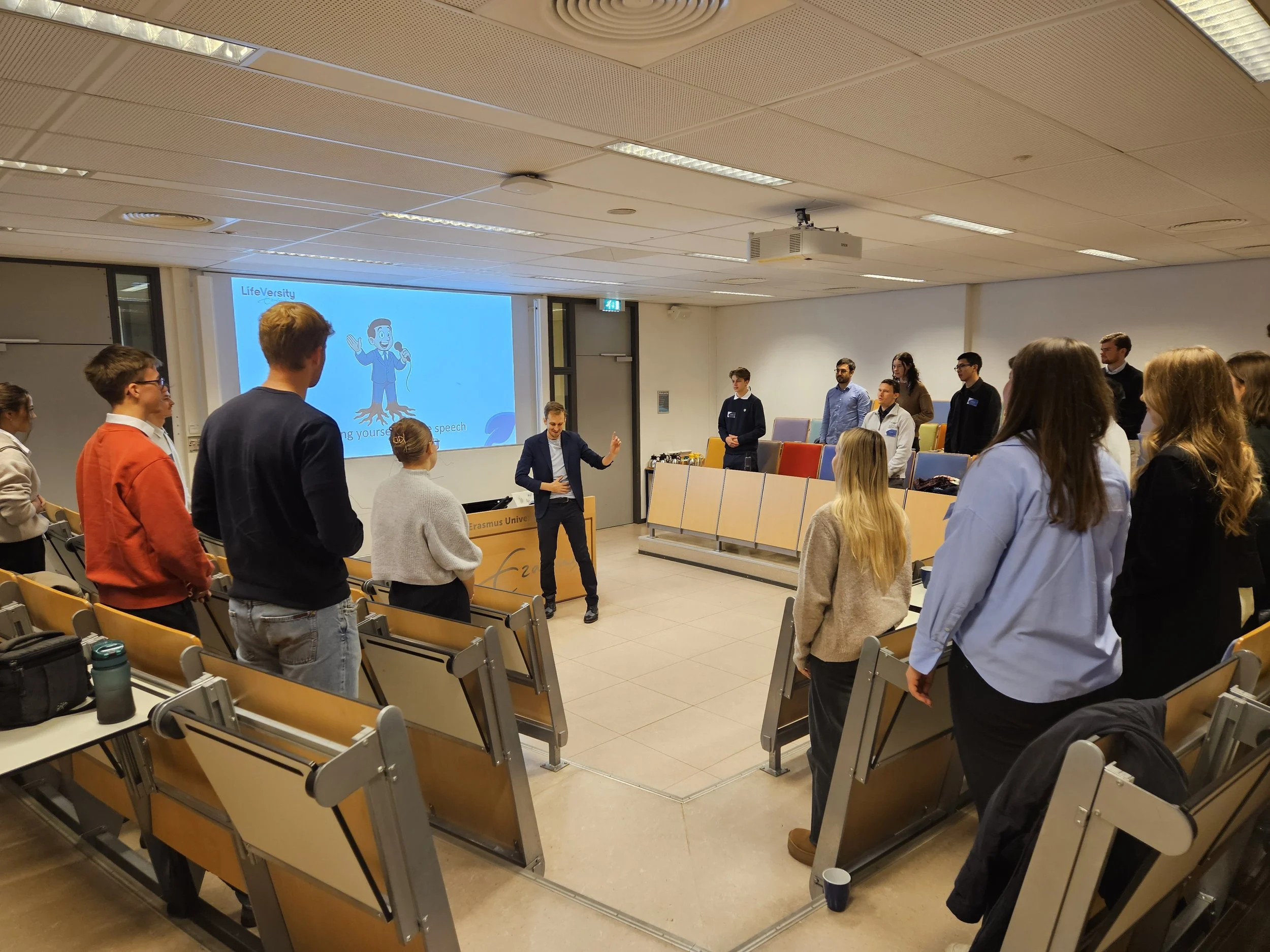 A group of people standing in a classroom or conference room, possibly for a presentation or workshop, with a man speaking at the front near a screen displaying a cartoon mascot with a microphone and the word 'LifeVersity'.