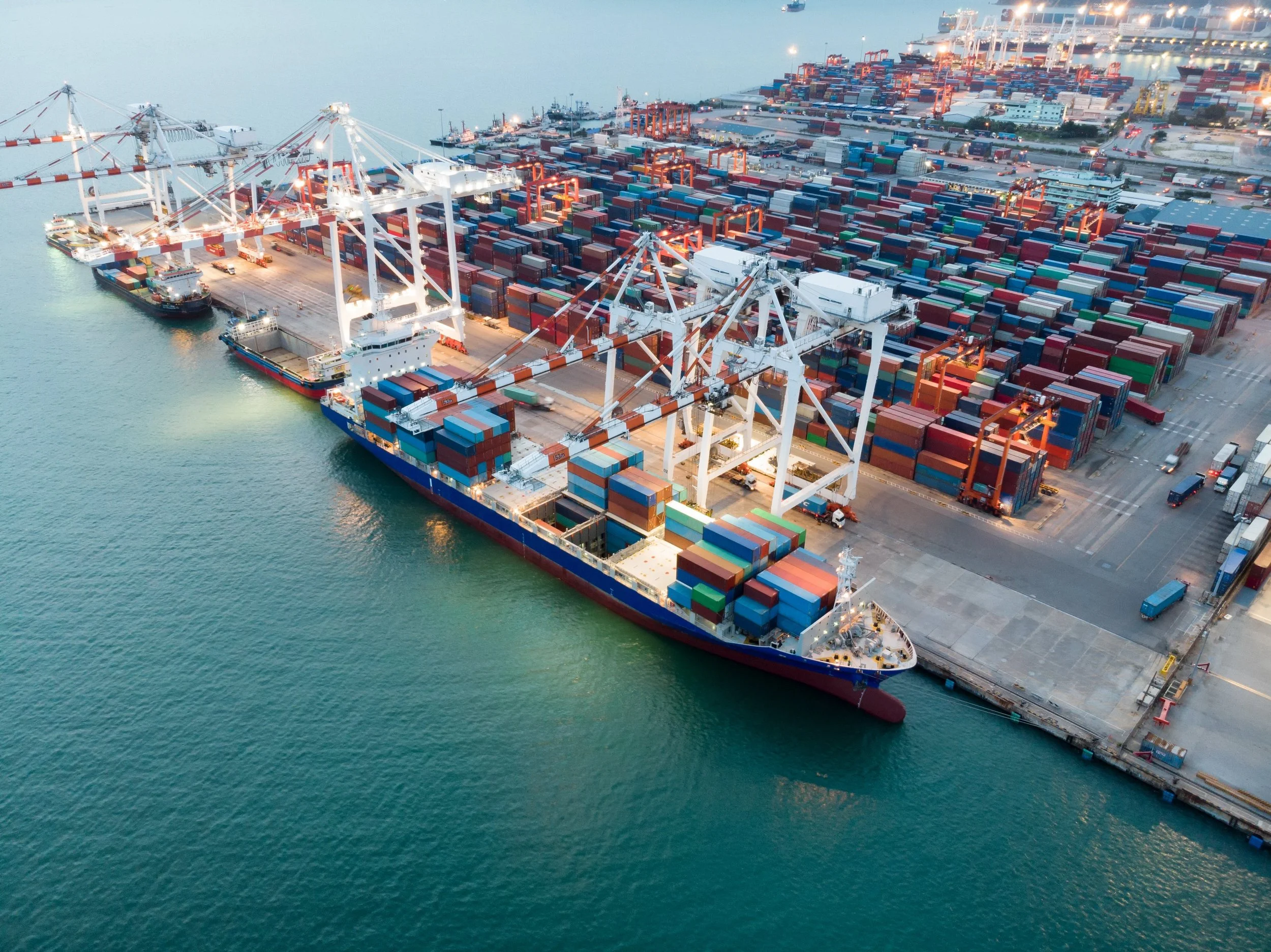 An aerial view of a busy shipping port with large cargo ships docked along the pier, surrounded by stacks of colorful shipping containers and towering crane equipment for loading and unloading containers.