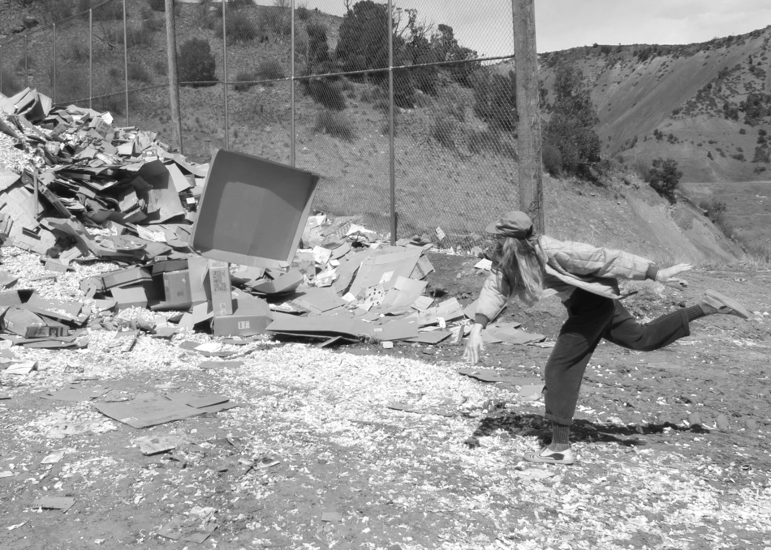 A person with long hair, wearing a cap, jacket, and pants, is in a dynamic pose throwing cardboard amidst a pile of cardboard and trash outdoors, with a fence and hills in the background.