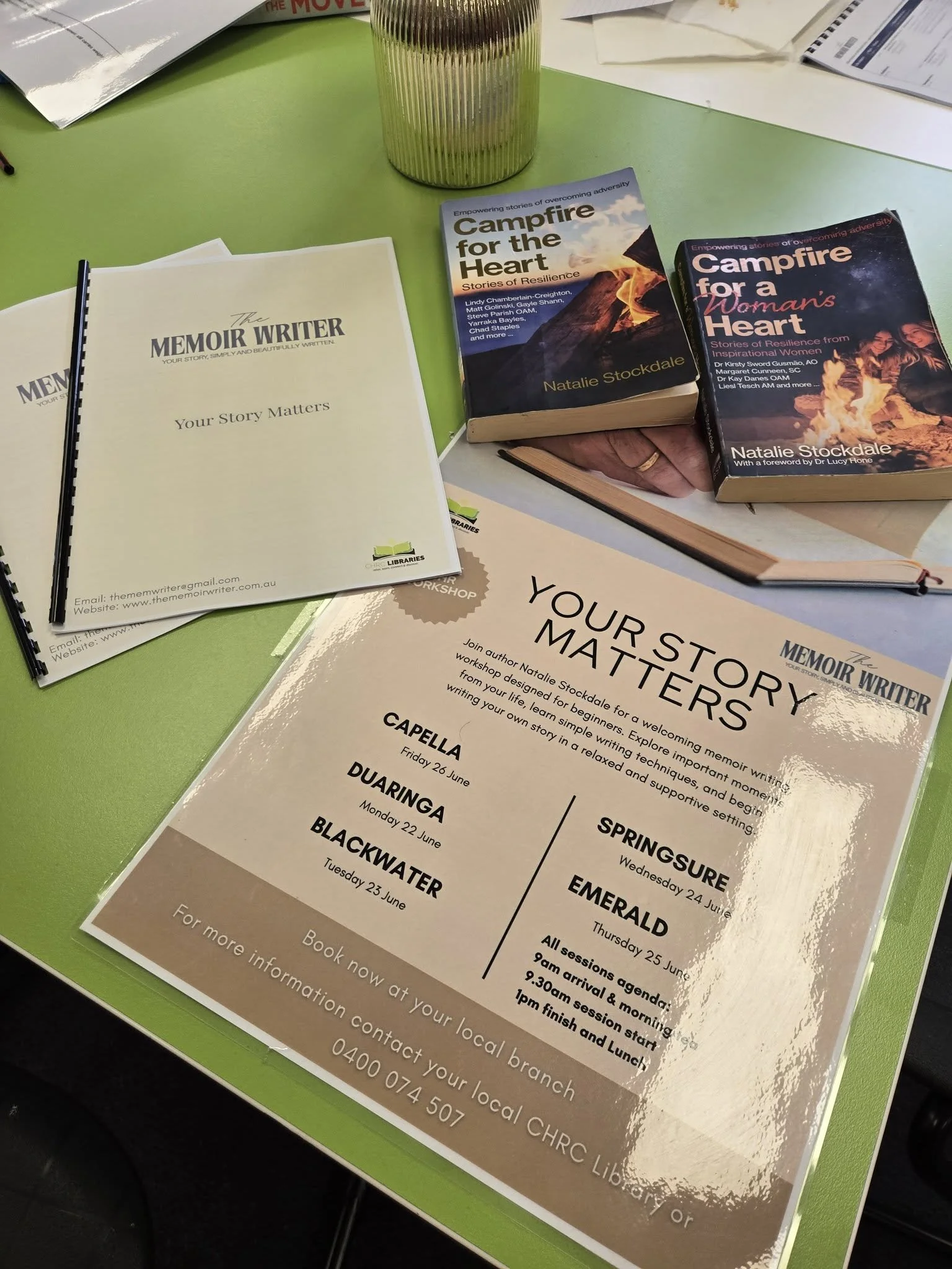 A pamphlet titled 'Your Story Matters' from The Memoir Writer, displaying contact information and website details. Next to it are two books titled 'Campfire for the Heart' and 'Campfire for a Woman's Heart' by Natalie Stockdale, placed on a green table. A transparent glass containing pens is also visible.